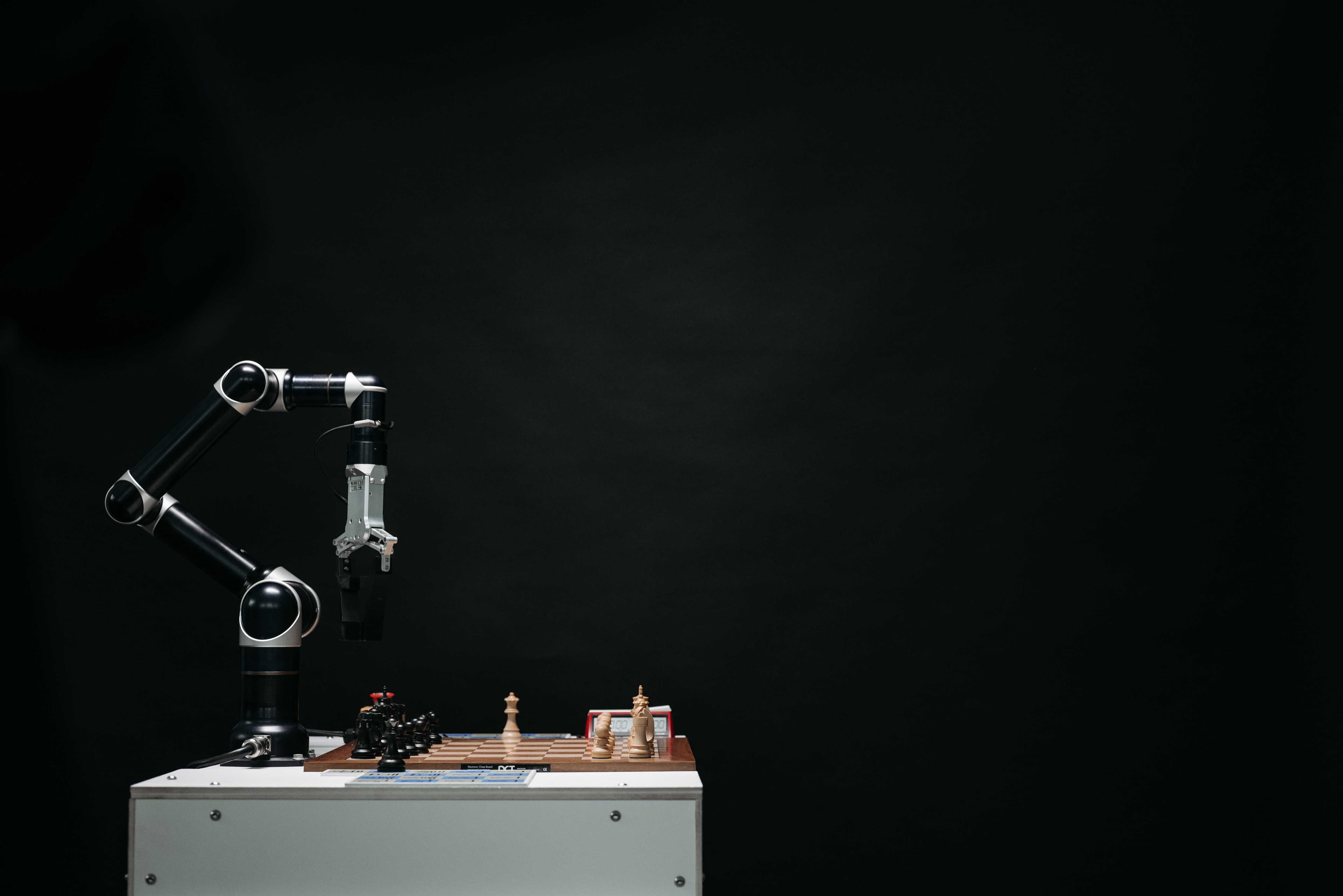 An AI robot arm that is playing chess on a wooden chess board on the brown side inside a dark room all on a white tabletop.