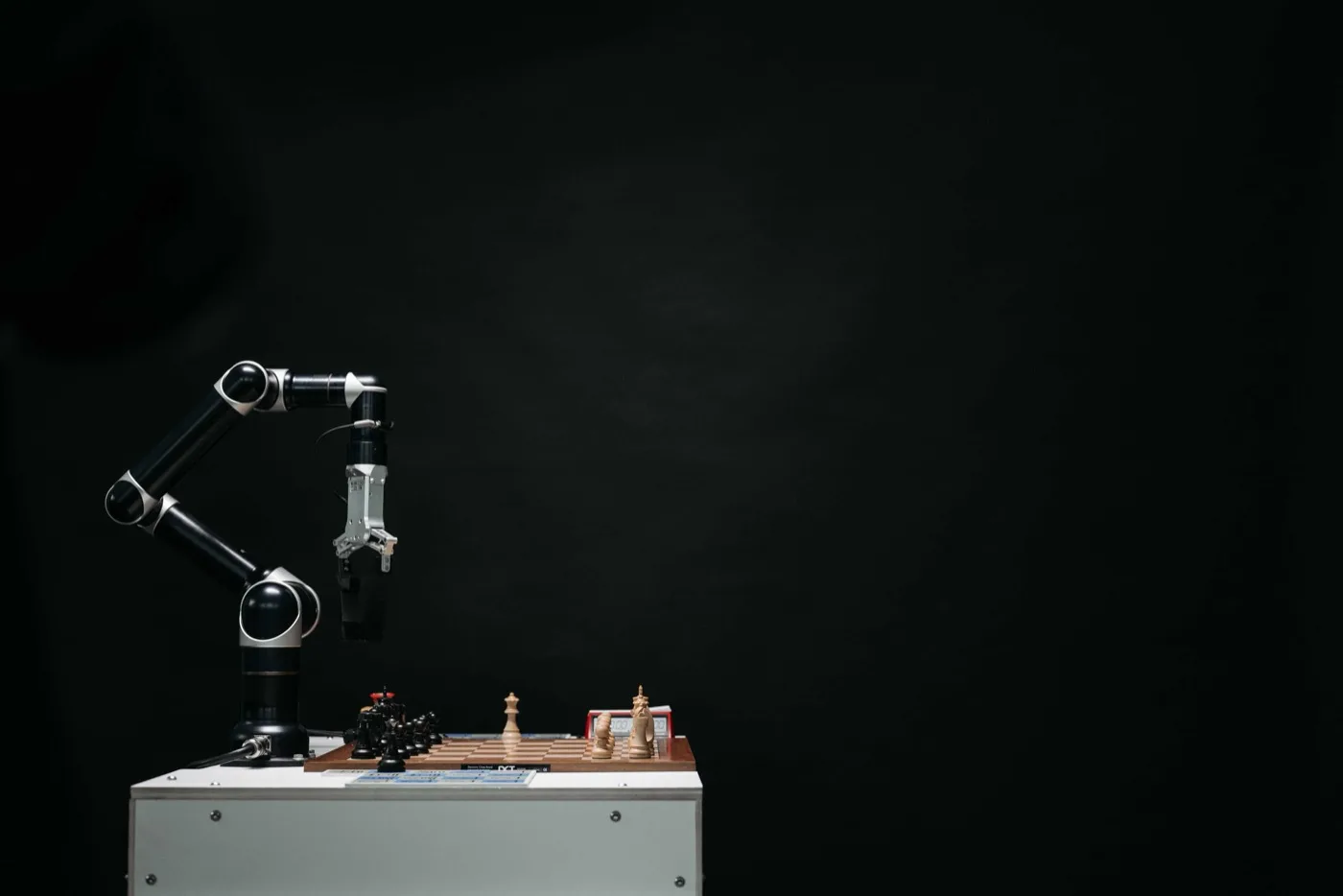 An AI robot arm that is playing chess on a wooden chess board on the brown side inside a dark room all on a white tabletop.