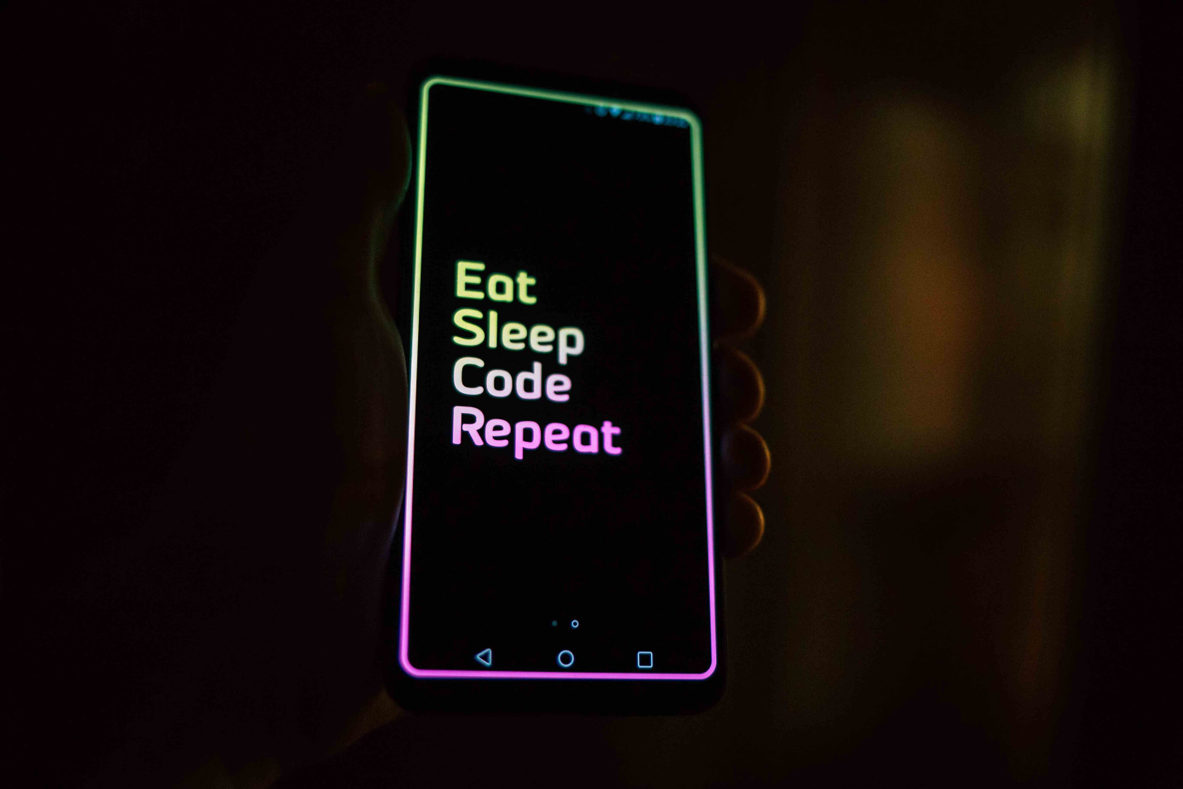 A smartphone that says Eat, Sleep, Code, Repeat, on it with a rainbow colored font in someone's hand surrounded by darkness.