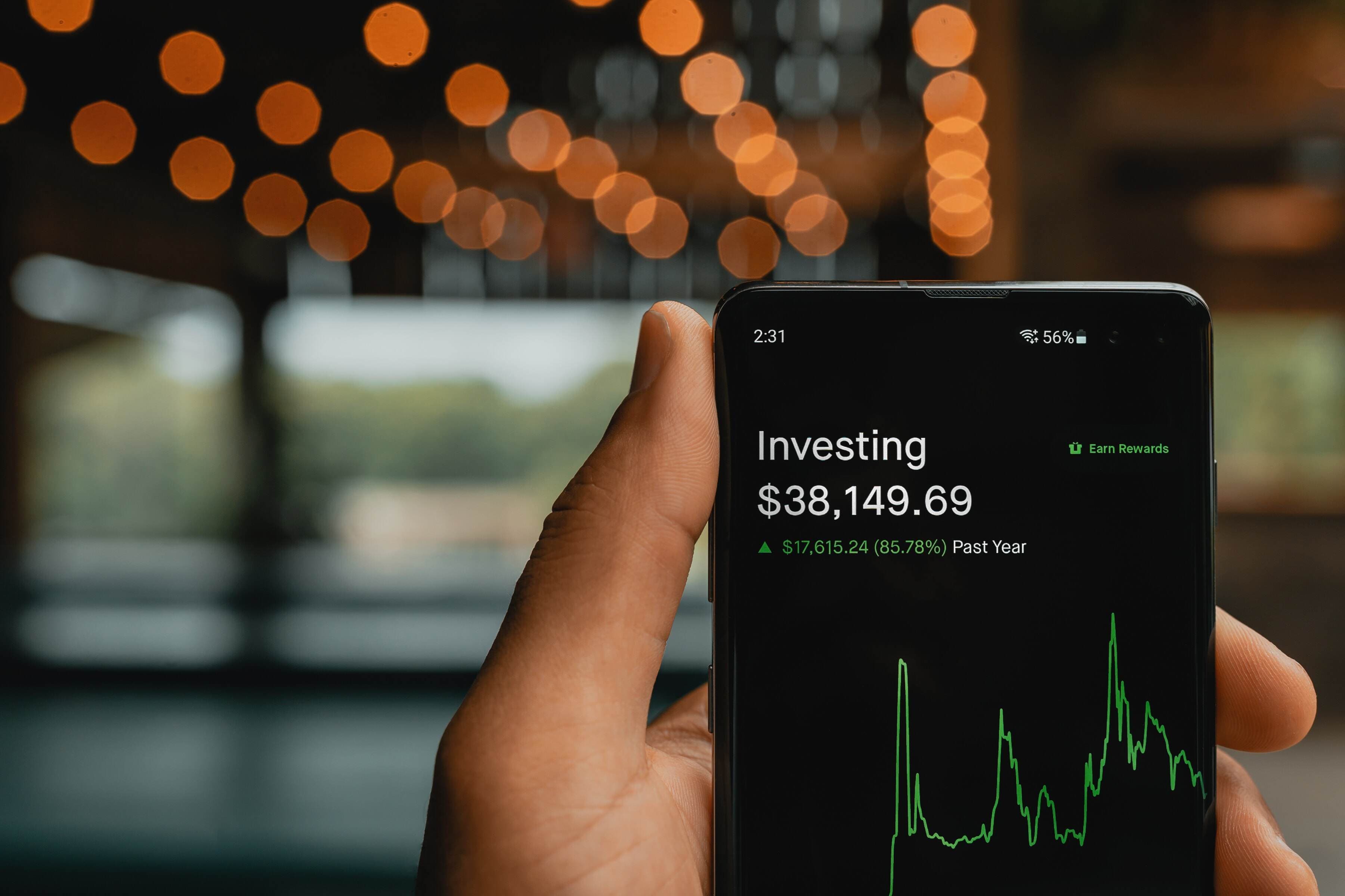 A smartphone in someone’s hand with a black screen that has an investing portfolio balance of $38,000 and a green line chart.