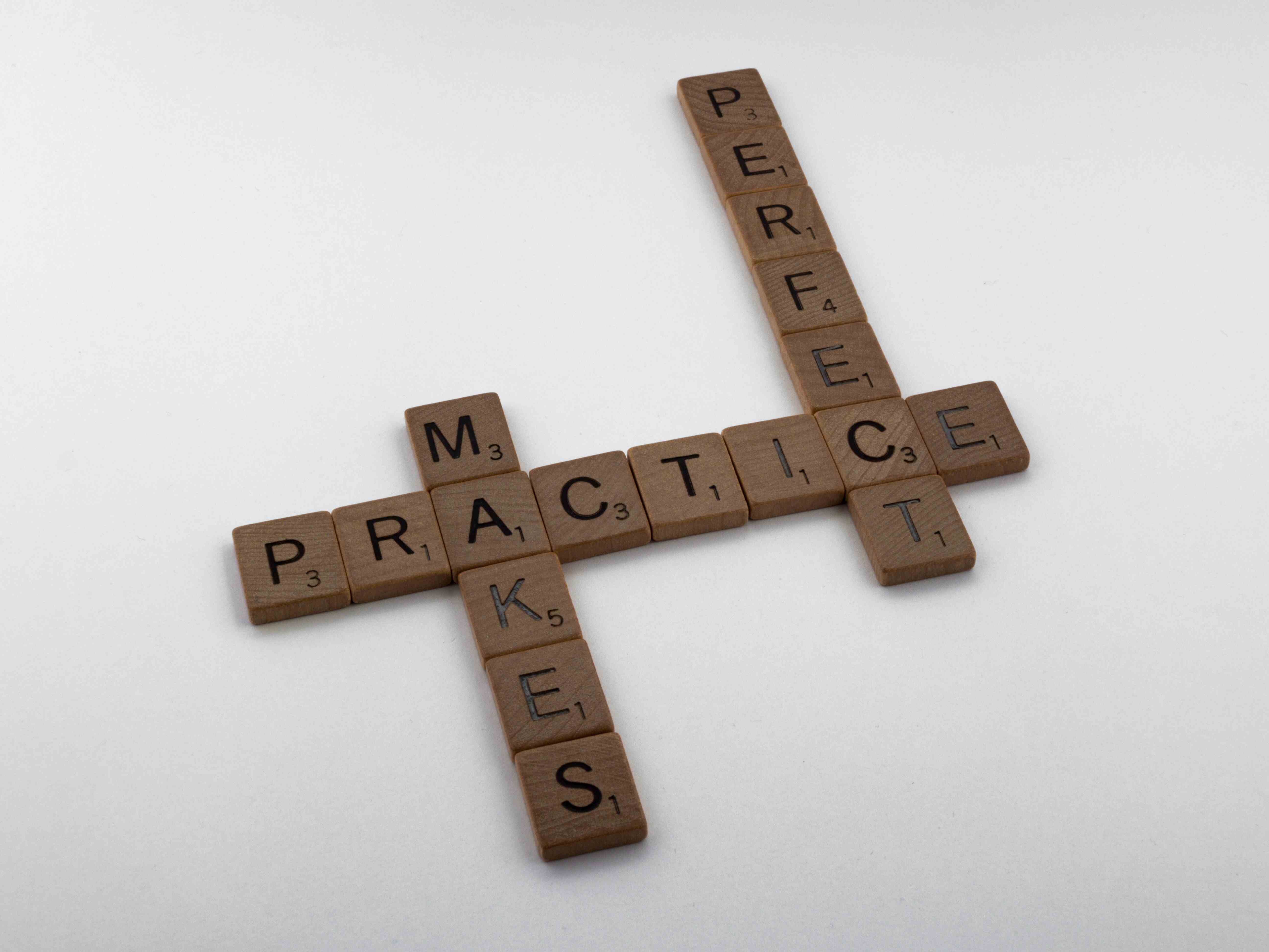 Dark wooden scrabble letters with black writing that write out practice makes perfect all connected together on a white surface.