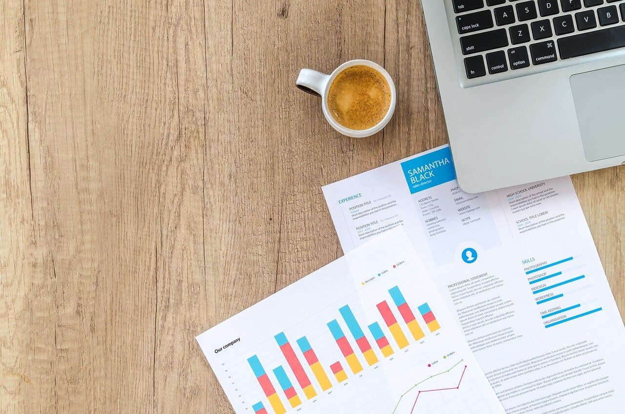 Two pieces of paper with multicolored graphs on them on top of a wooden table next to a cup of coffee and a Macbook Laptop.