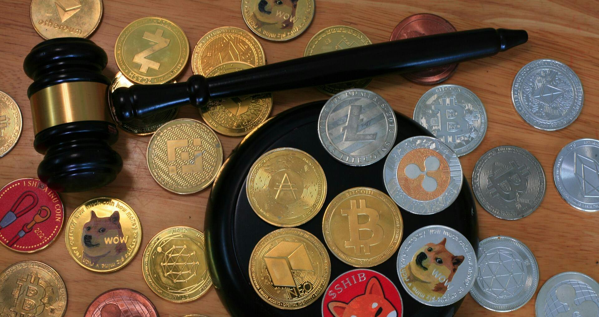 Twenty-five different shiny crypto coins that are both gold and silver on a table with a wooden gavel on the table with them.