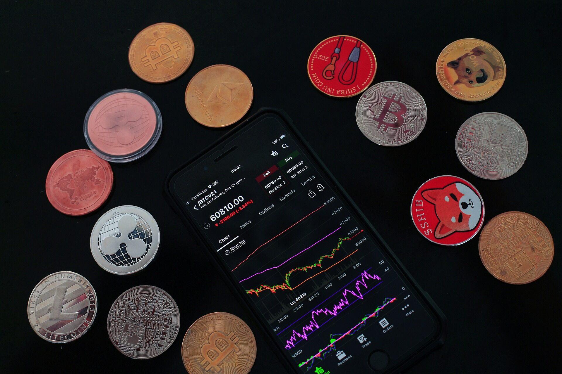 Fourteen different crypto shiny coins in the colors of gold, silver, and red next to an Apple iPhone that is showing the market.