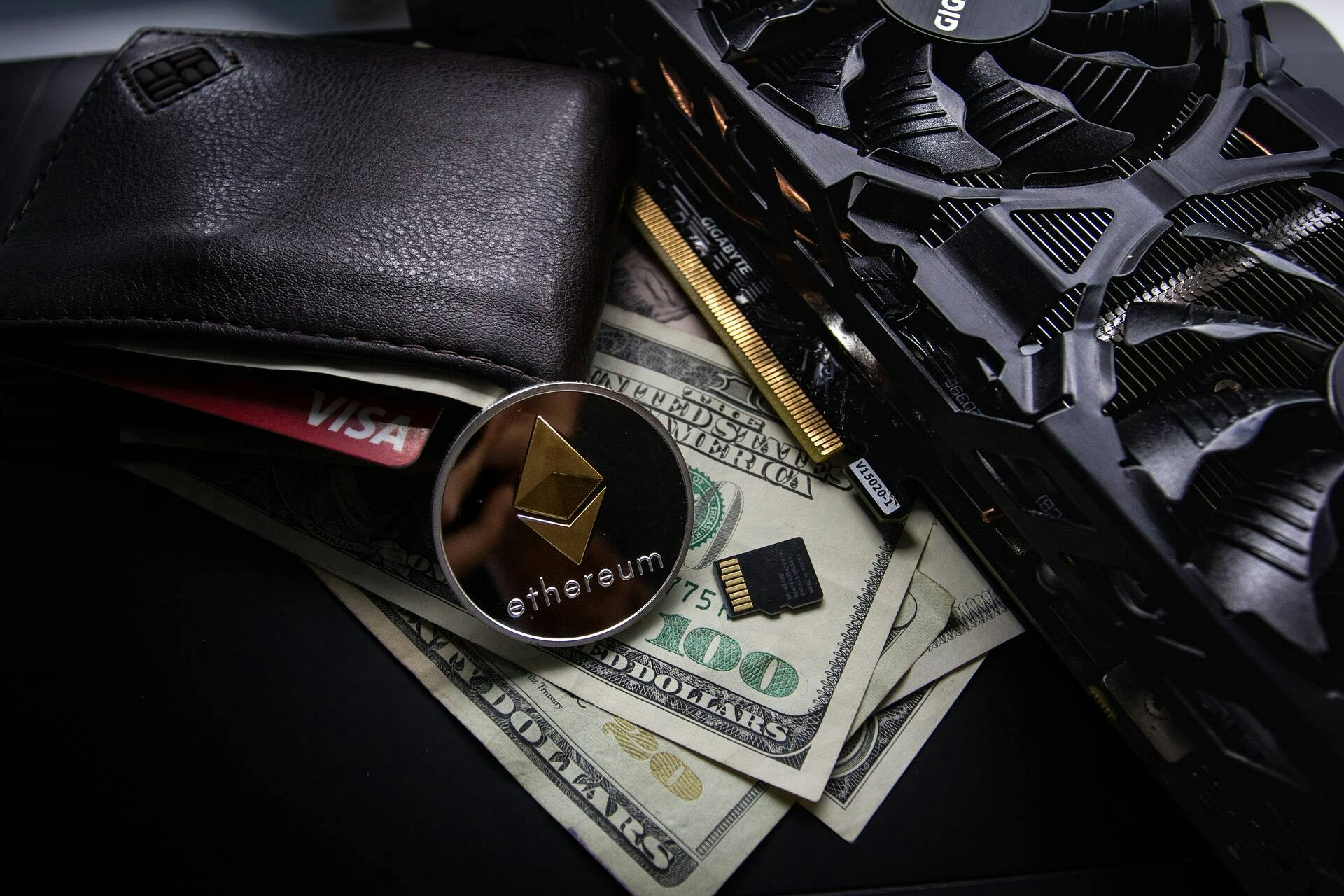 A shiny metal Ethereum crypto coin with a leather wallet next to it containing a credit card all on to pof a stack of dollars.