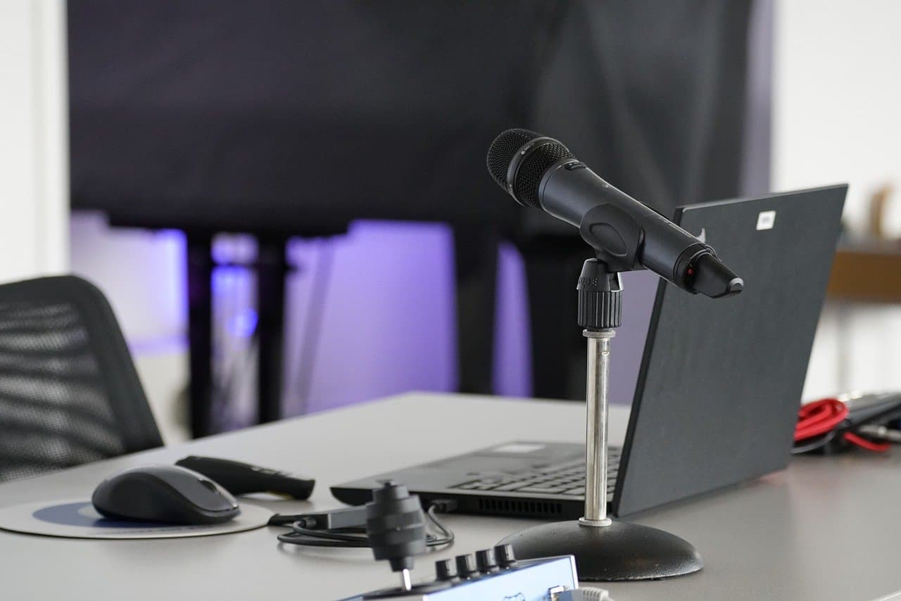 A black laptop on a white table next to a black microphone and a black mouse with a black chair sitting underneath the table.