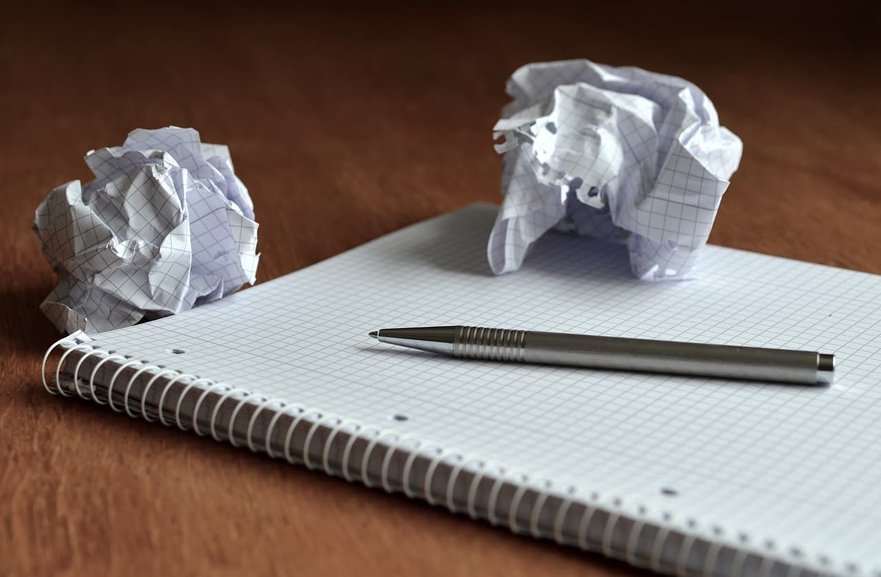 A white graphing paper notebook with a white spiral and silver metal pen resting on top next to two balled-up pieces of paper.