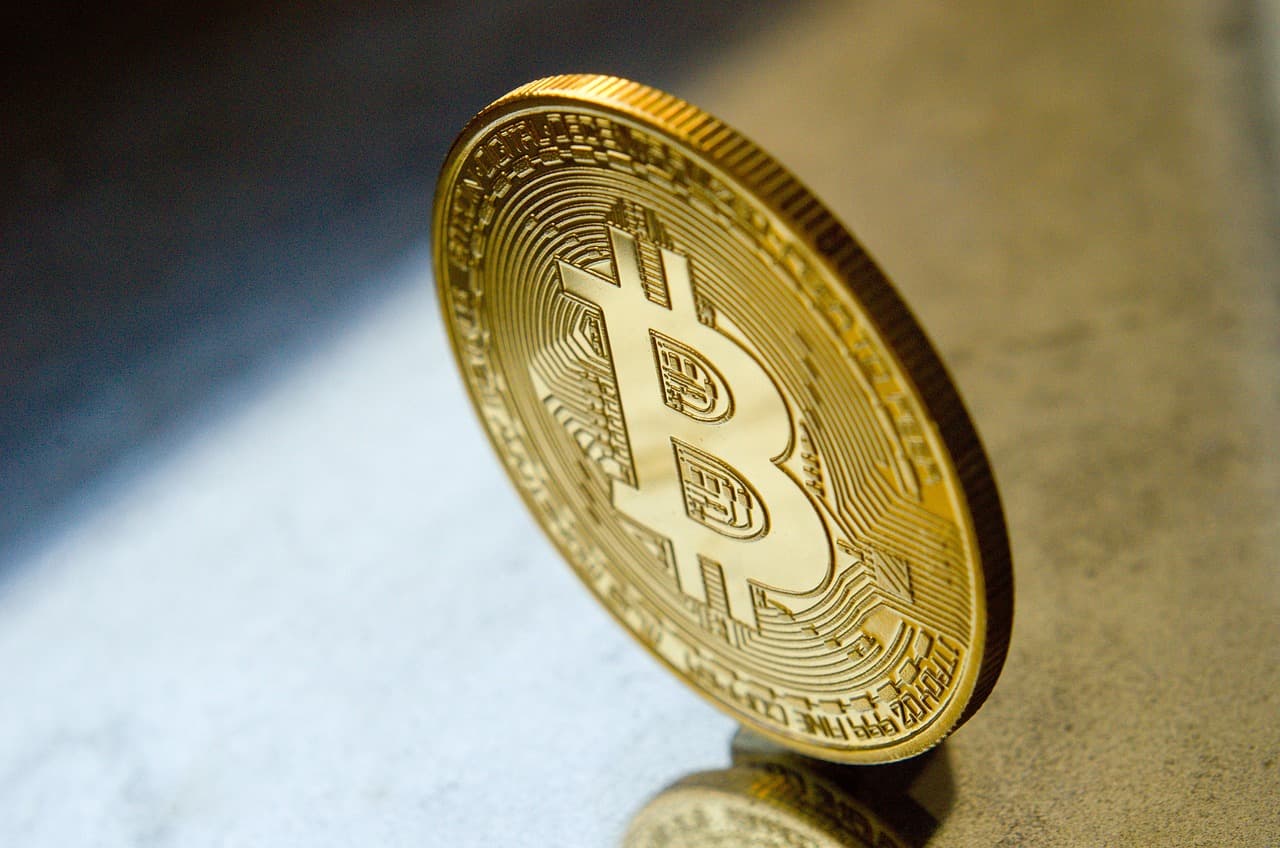 A golden coin with the Bitcoin logo imprinted onto it is sitting on its side on top of a grey-colored stone polished surface.