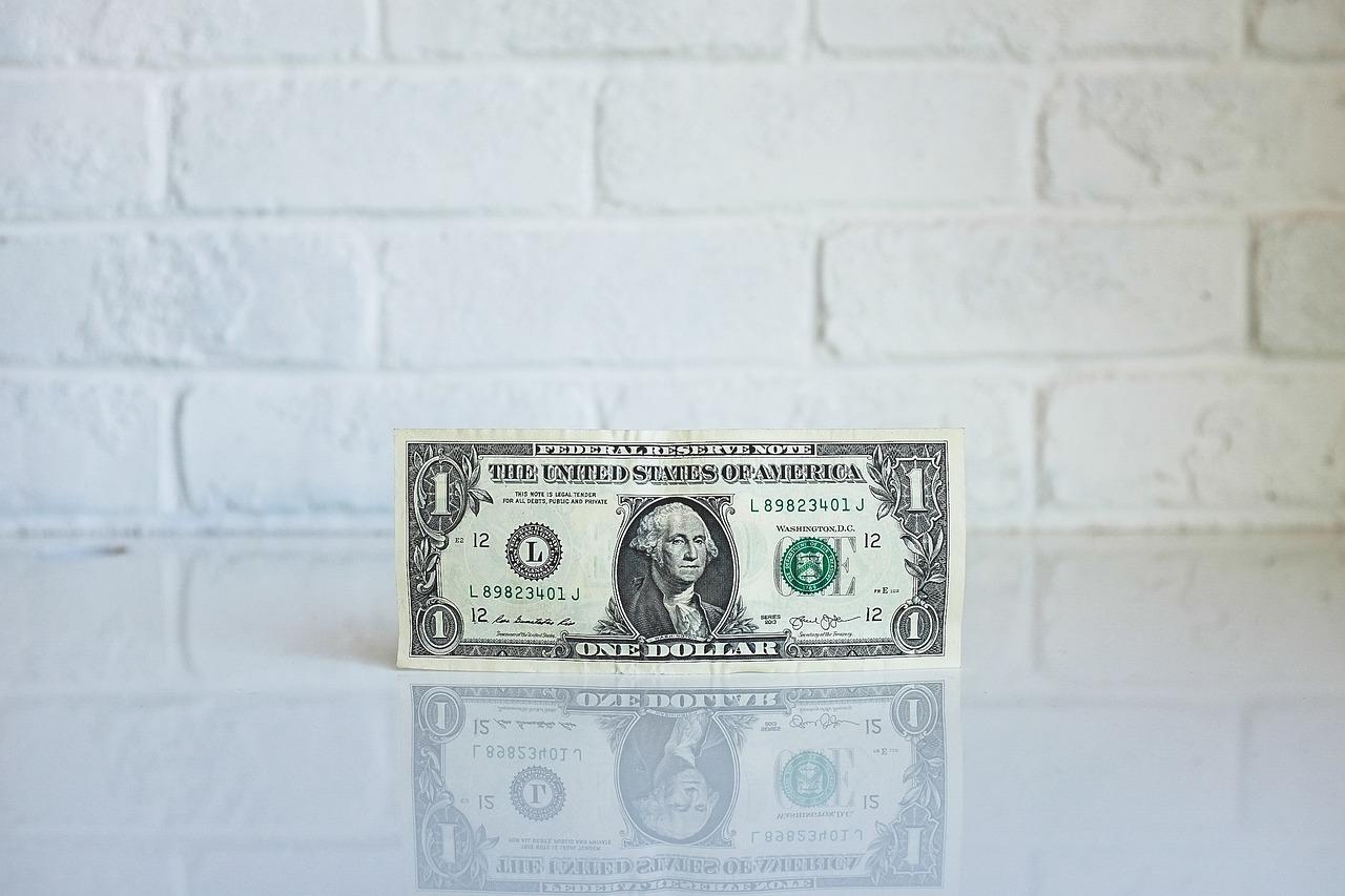 A one dollar bill standing up on its edge with the face side forward in front of a brick wall that has been painted white.