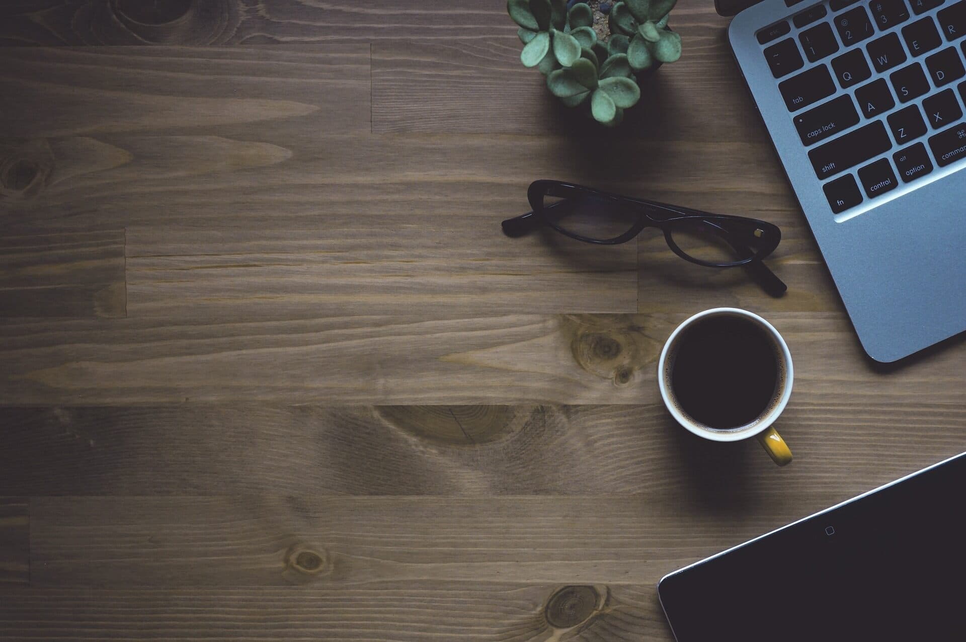 A cup of coffee with a small green plant next to black glasses and an open Apple Macbook laptop all on a dark wooden table.