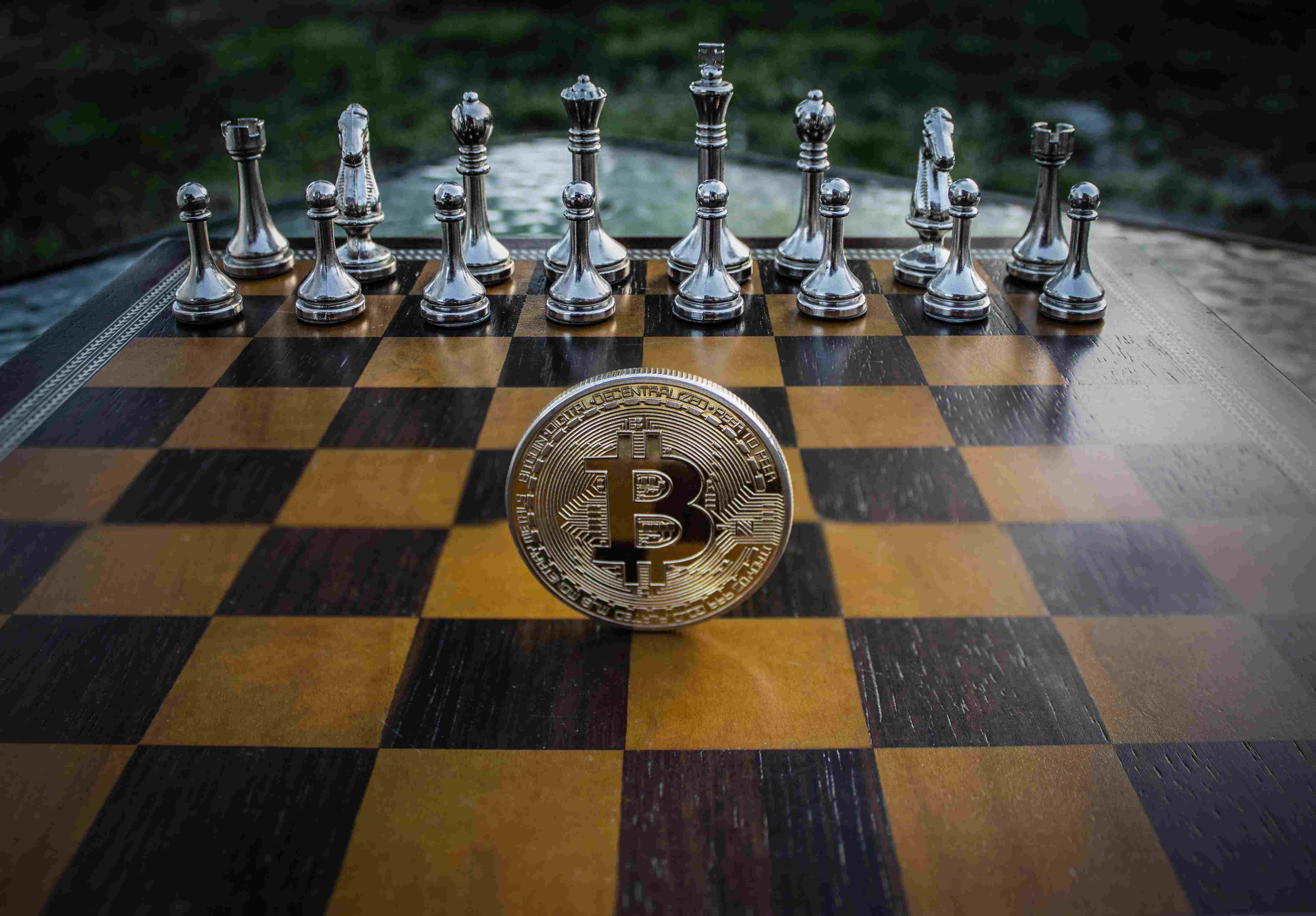 A Bitcoin sitting on top of a wooden chess table with the white pieces all lined up in the background in an outdoor setting.