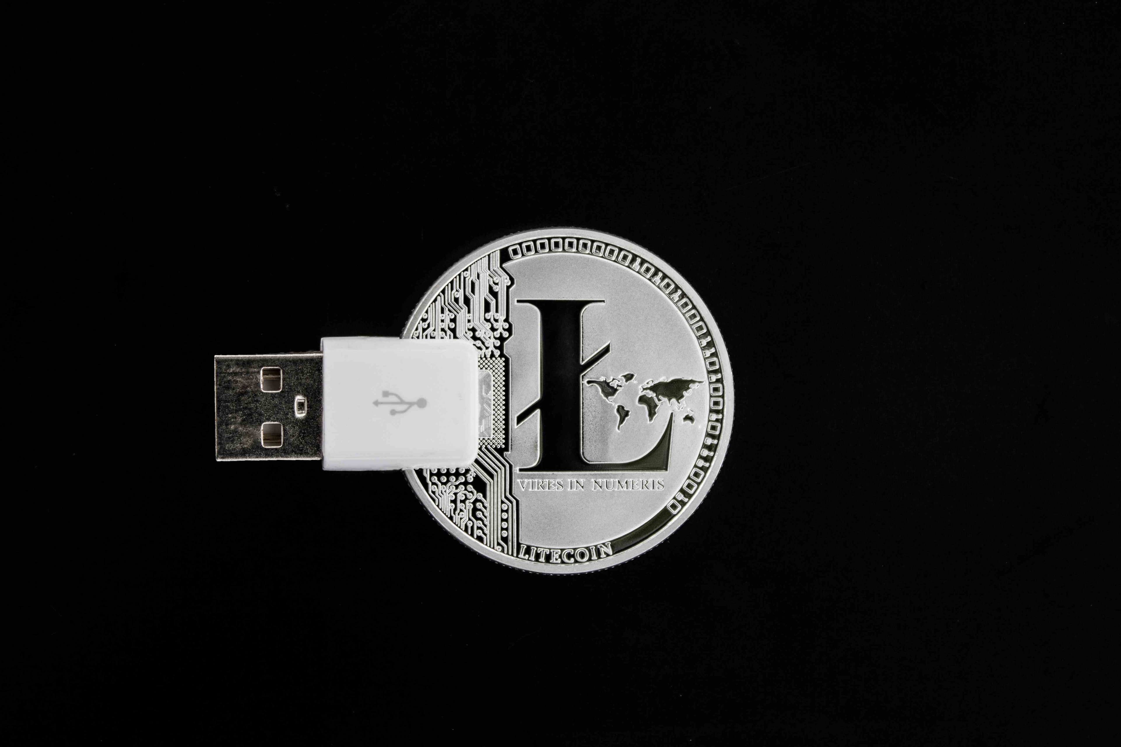 A silver Litecoin with a white USB male end resting on top of the coin showing the USB symbol all sitting on a black surface.