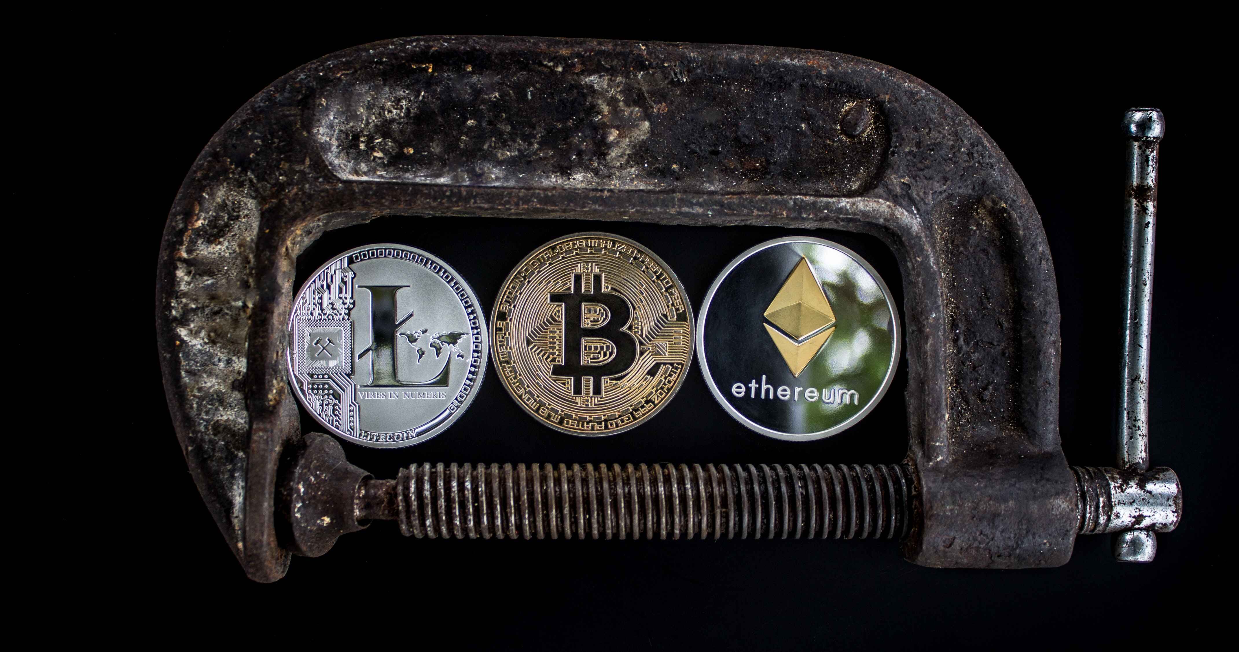 Litecoin, Ethereum, and Bitcoin are all sitting inside a brown and rusty vice grip that is in front of a black background.