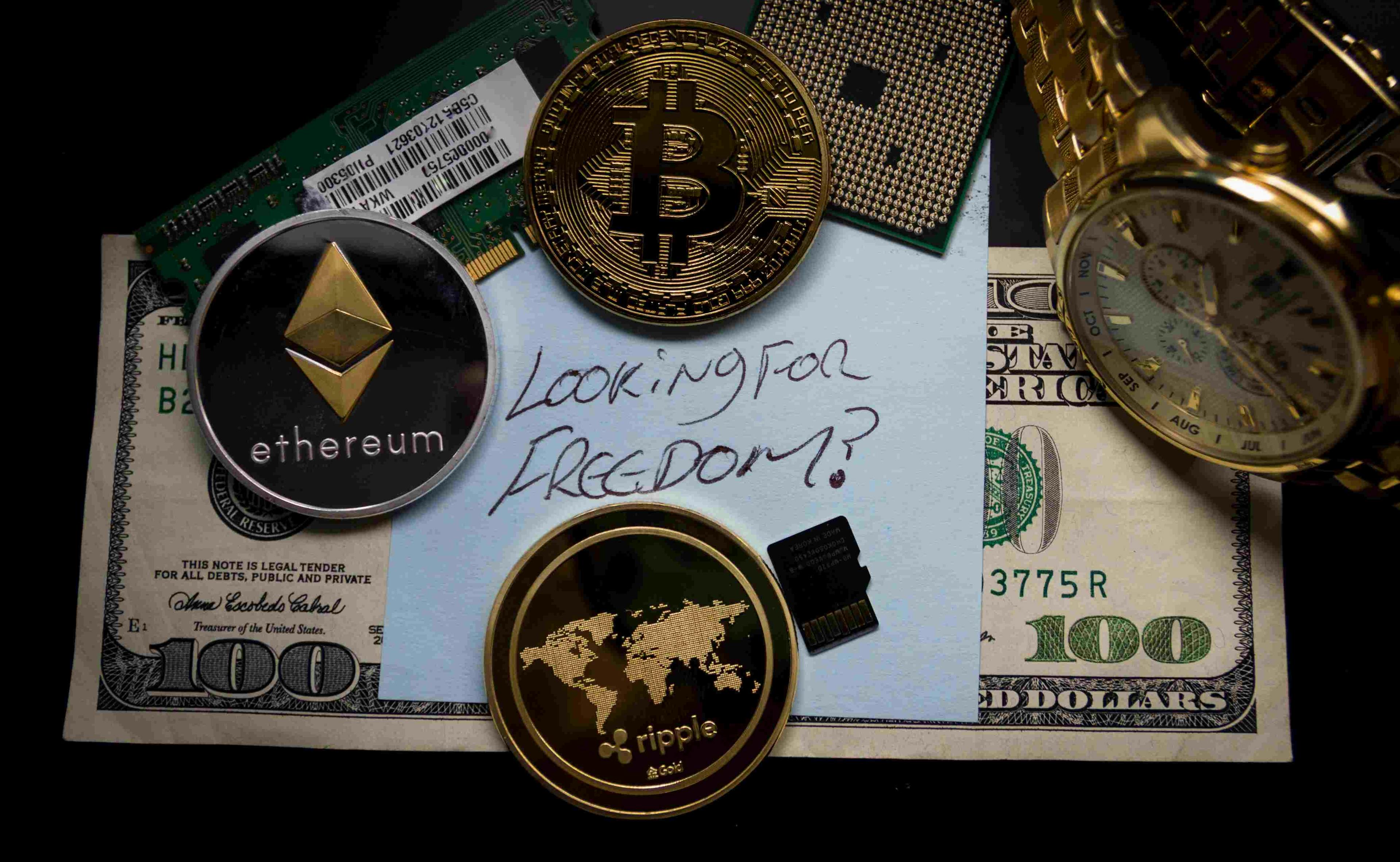 Three cryptocurrency coins resting on top of a one hundred dollar bill with a piece of paper on top and a gold watch next to it.