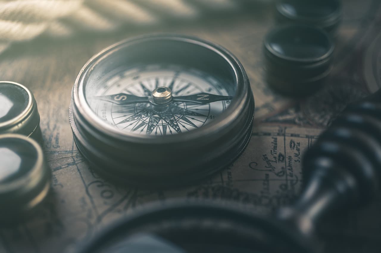 A brown rustic metal compass next to small and large magnifying glasses all on top of a lightly colored map with black lines.