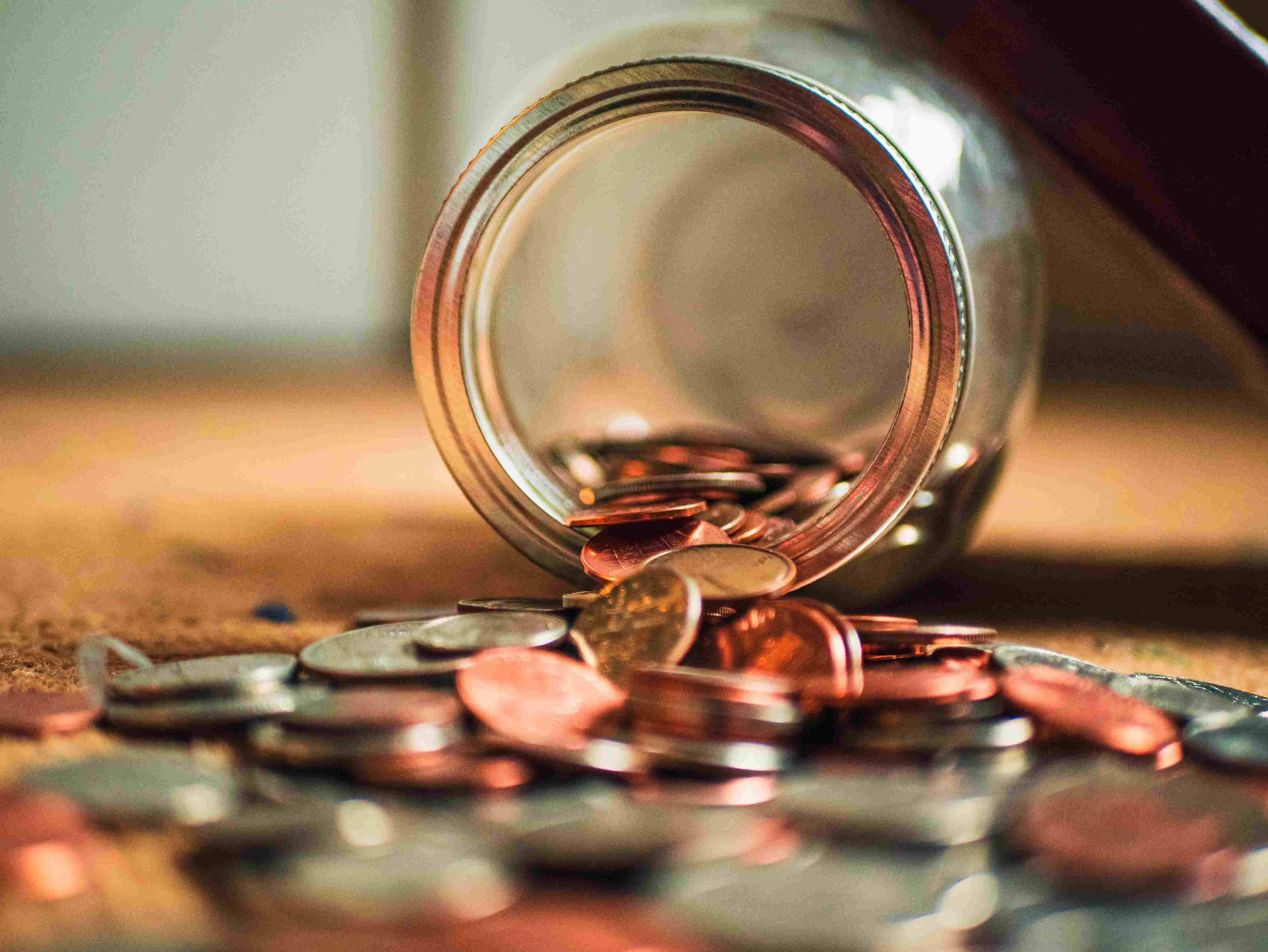 A tipped-over glass jar that is holding a large number of bronze coins with some spilled out onto a light brown surface.