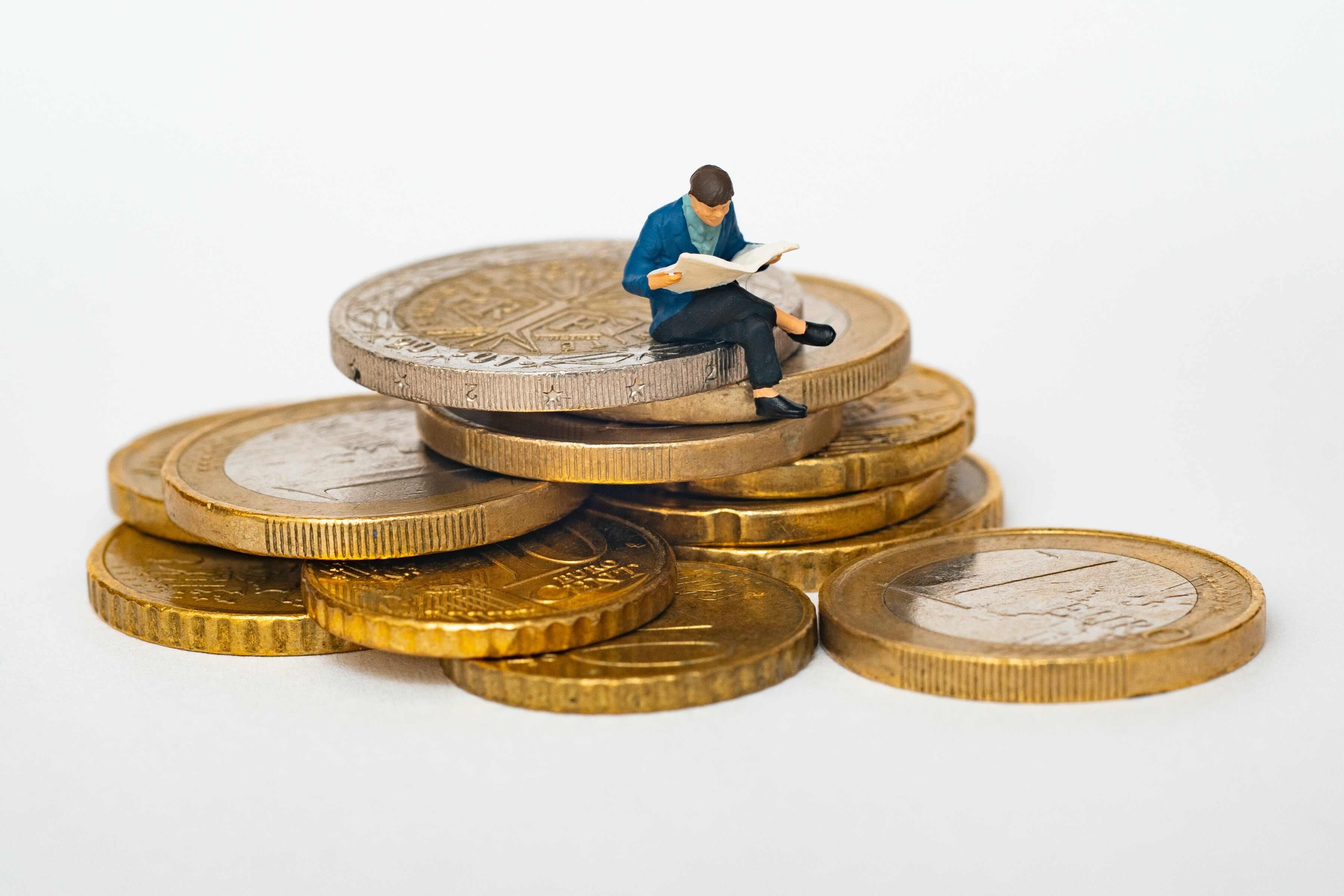 A tiny clay man in a blue jacket and brown hair reading a book sitting on a pile of eleven foreign coins with gold coatings.