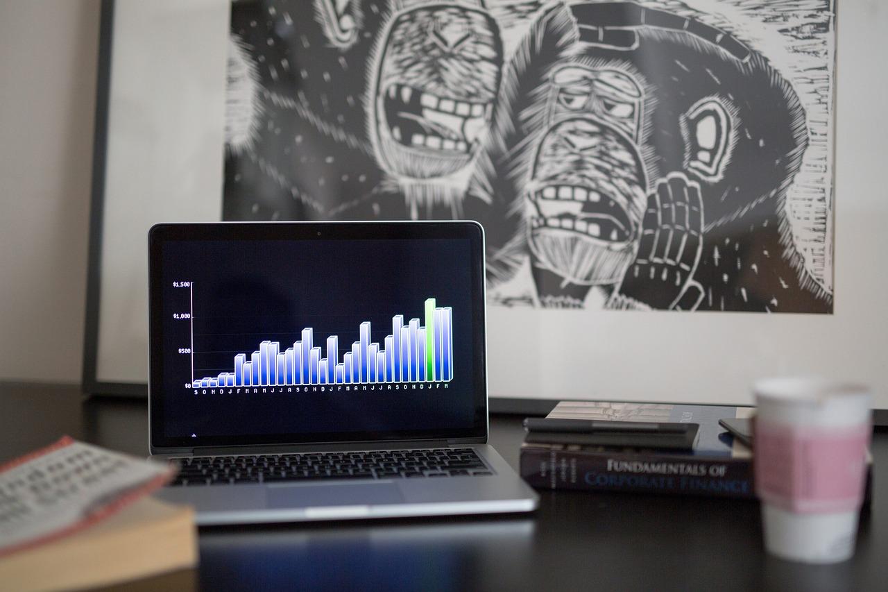 A silver Apple Macbook with a blue and white bar chart on it on a black desk with two books and a cup of coffee next to it.