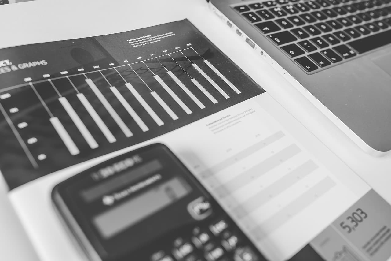 A black and white bar chart on a piece of white paper in a spiral notebook with a thick calculator on top of it next to a laptop.