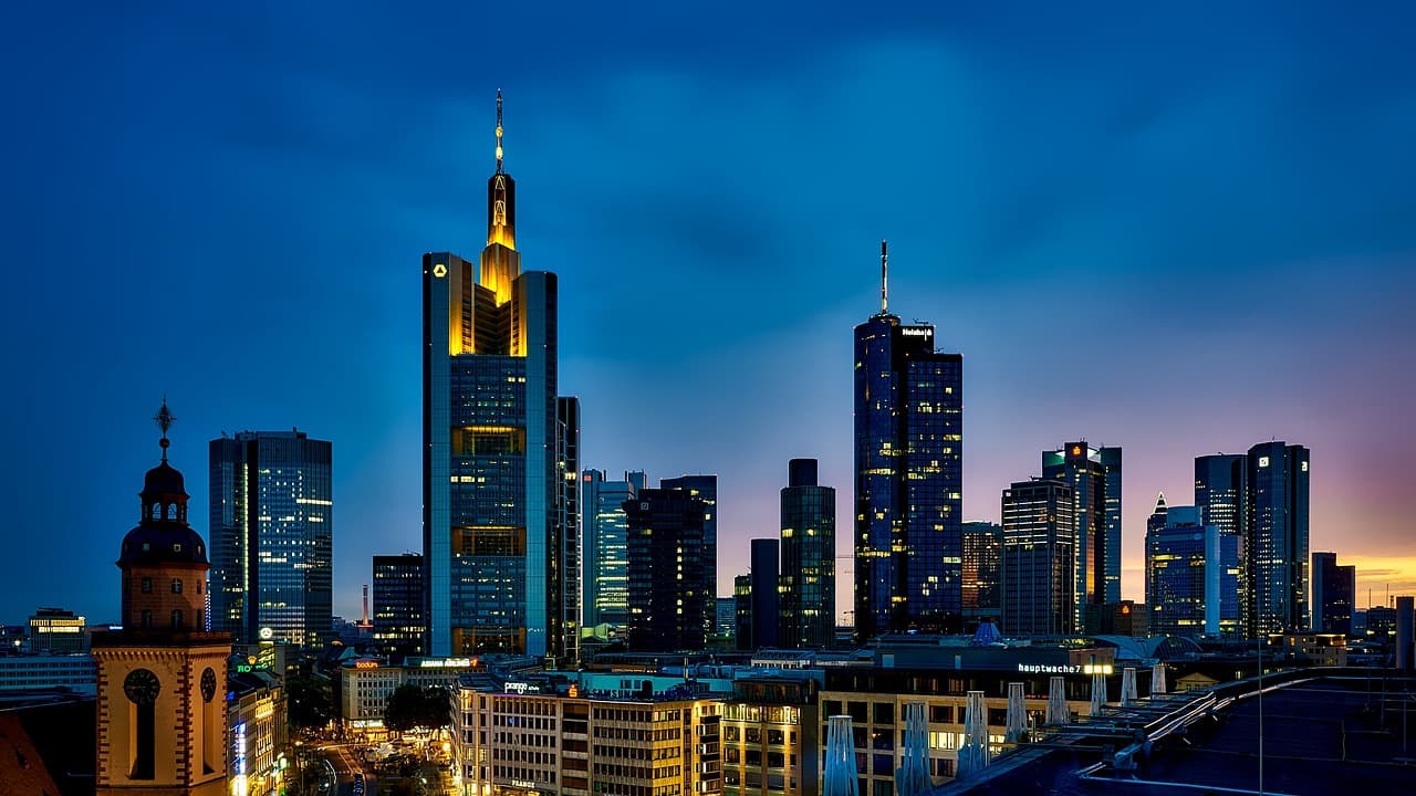 A blue skyline filled with tall buildings in black with yellow lights in their windows with the sunset shining behind them.