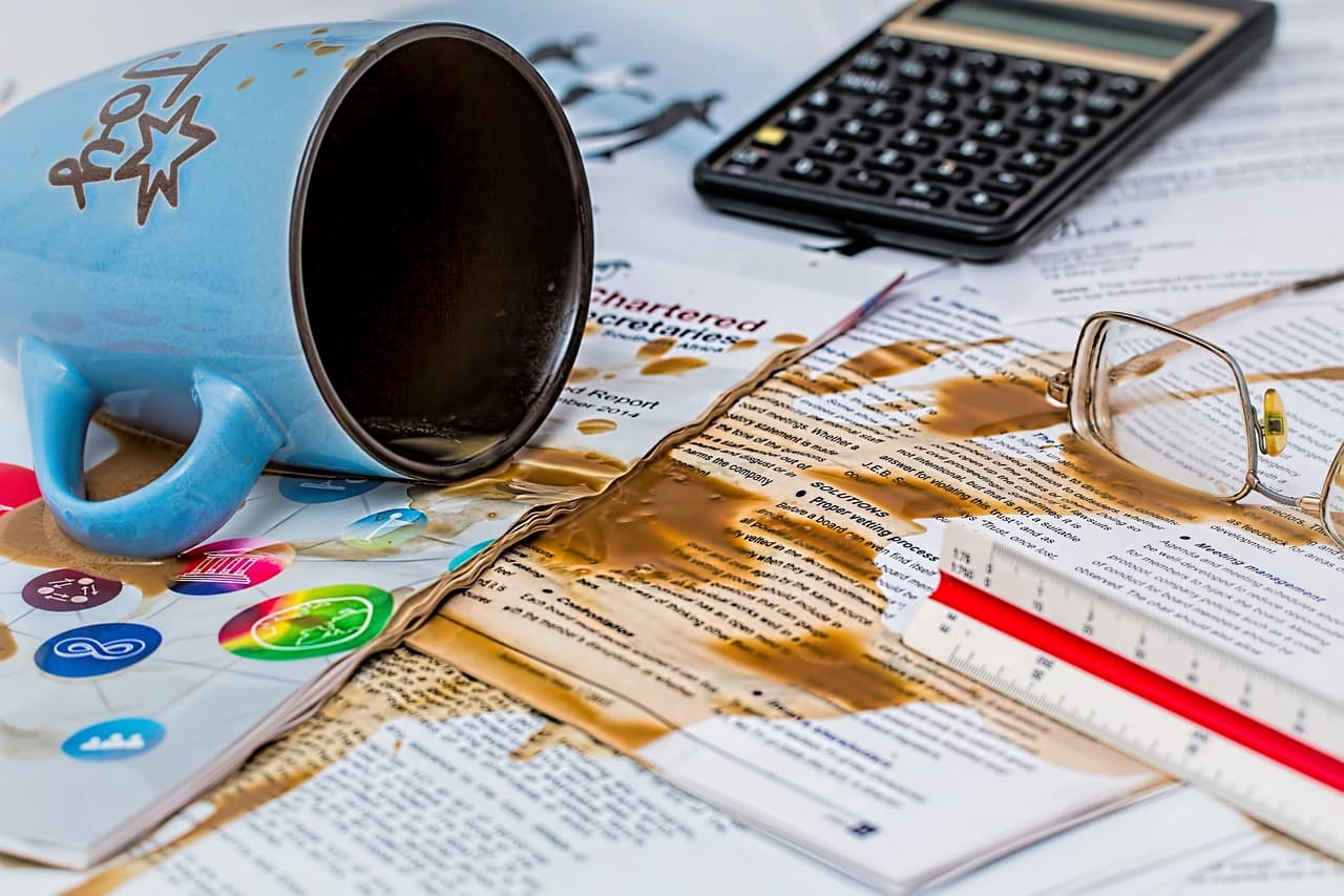 A blue cup of coffee that is tipped over spilling the brown coffee all over a white piece of paper with numbers and words on it