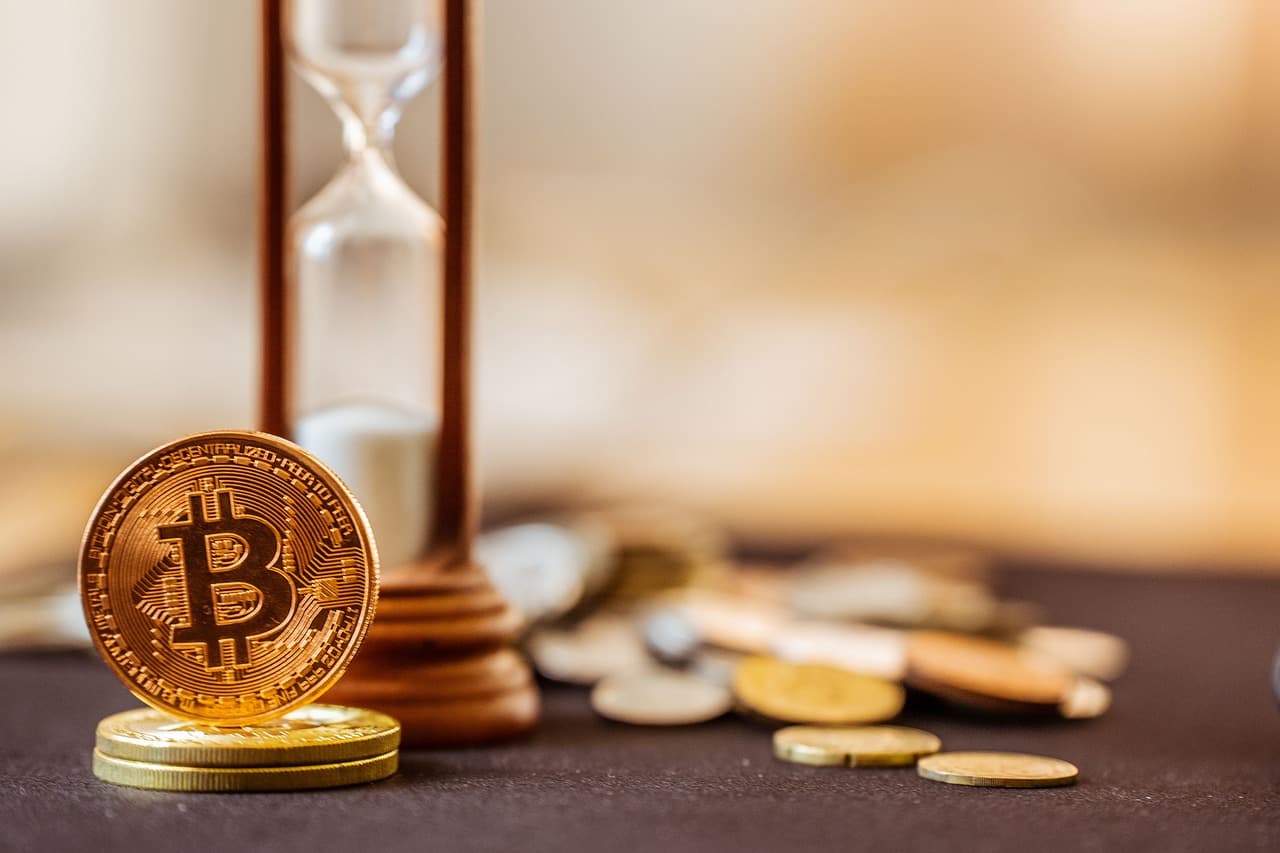 A golden coin on top of two other coins with the Bitcoin logo on the front of it in front of a wooden hourglass and other coins.