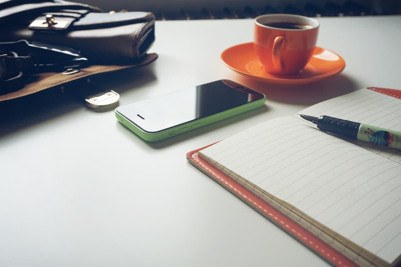 A notepad and a fountain pen on it for freelance work next to a blue smartphone and an orange cup on coffee on an orange plate.