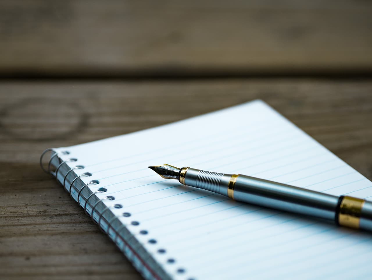 A white lined notebook with a silver fountain with bronze accents resting on it all on top of a dark brown wooden picnic table.