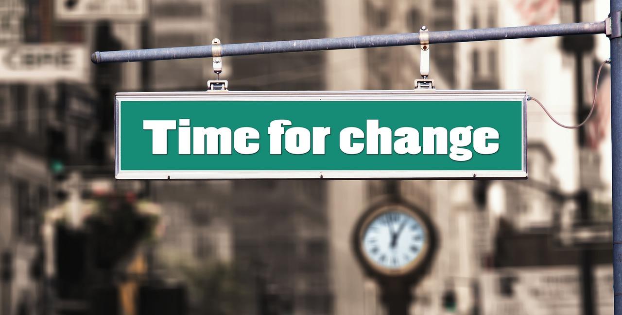 A green sign on a traffic light that says time for a change in white letters with a city in the background over a busy road.