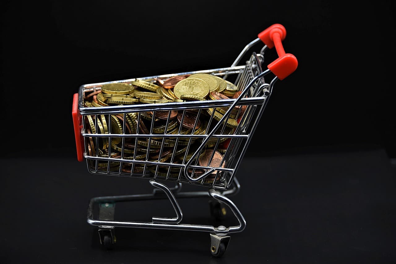 A mini shopping cart with a red handlebar that has multiple golden coins sitting inside of it on top of a black surface.