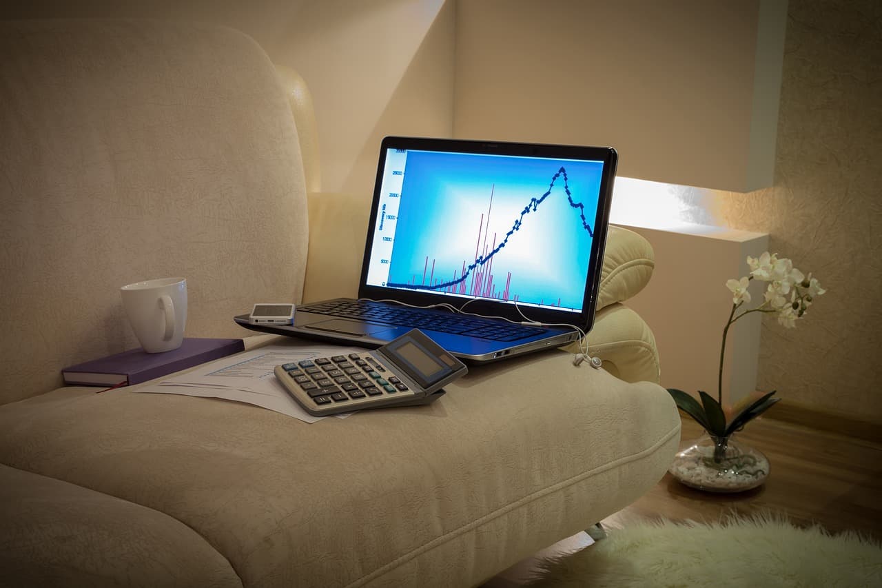 A dark blue chart on a blue screen of a silver Macbook on top of a tan couch next to a black notebook and a white cup of coffee.