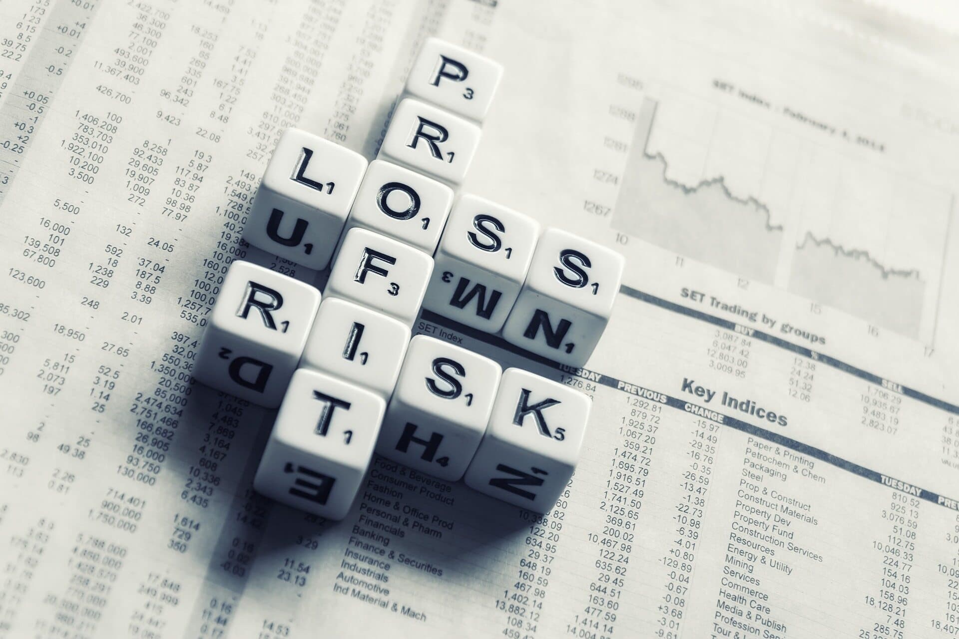 The words profit, loss, and risk, spelled out on a crossword section of a newspaper using white dice with letters in black.