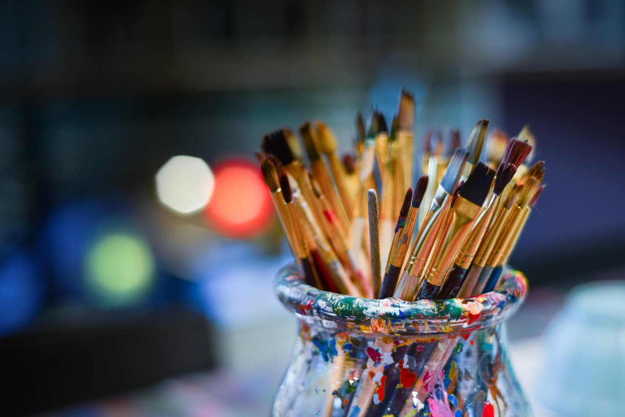 A glass cup that has multiple varying colors of paint on the outside of it with many differently colored paint brushes inside.