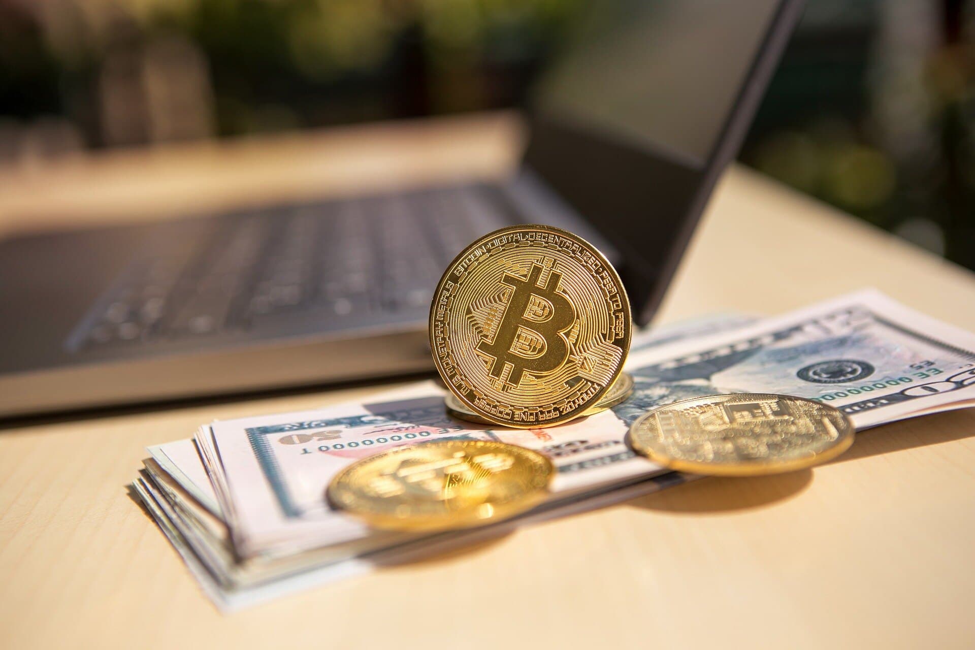 A golden bitcoin crypto coin sitting up on top of multiple hundred dollar bills with two other coins nearby and a macbook.