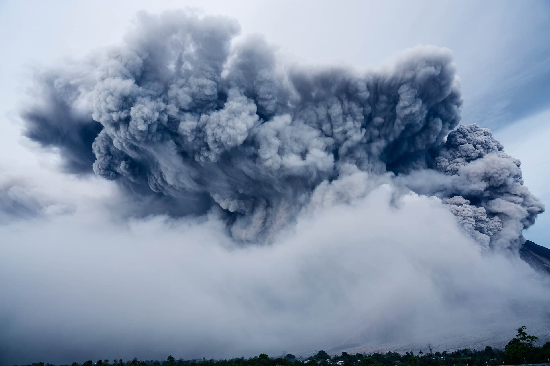 A large cloud of dark gray smoke with streaks in it surrounded by clouds that is erupting from a large rocky and sandy mountain.