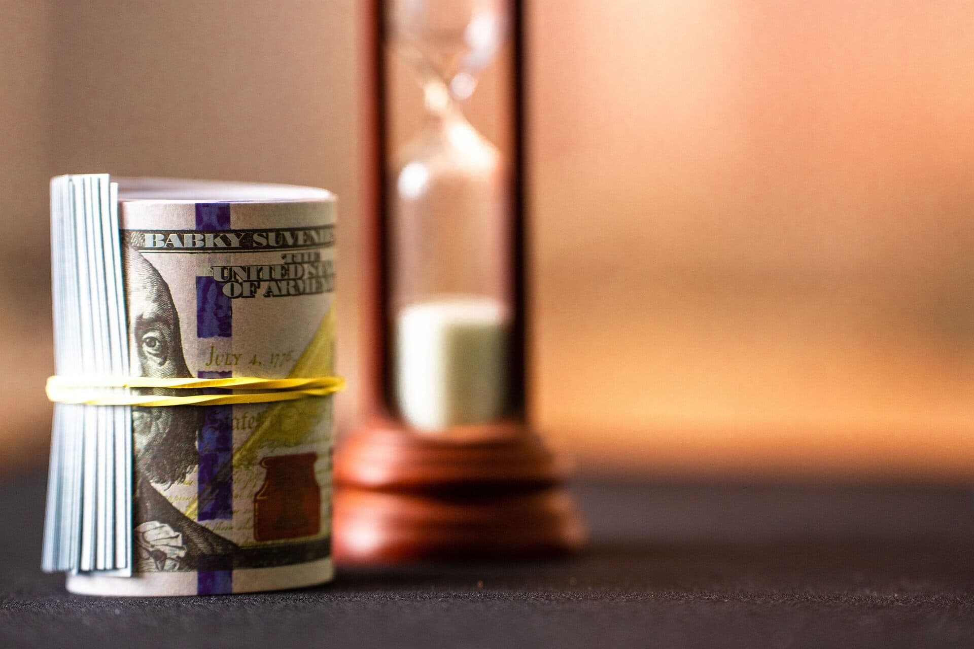 Stack of hundred dollar bills rolled up with a rubber band around them with a wooden hourglass in the background with white sand.