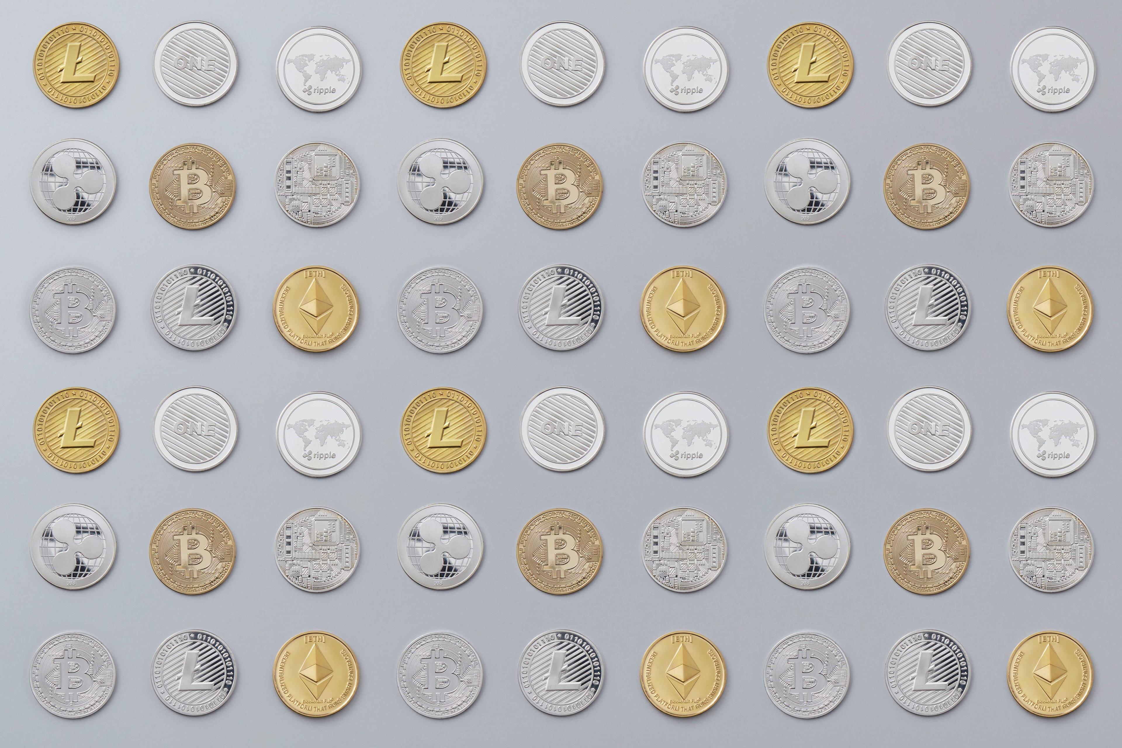 Fifty-four golden, bronze, and silver cryptocurrency coins all sitting out on a dark grey surface evenly separated and flat.