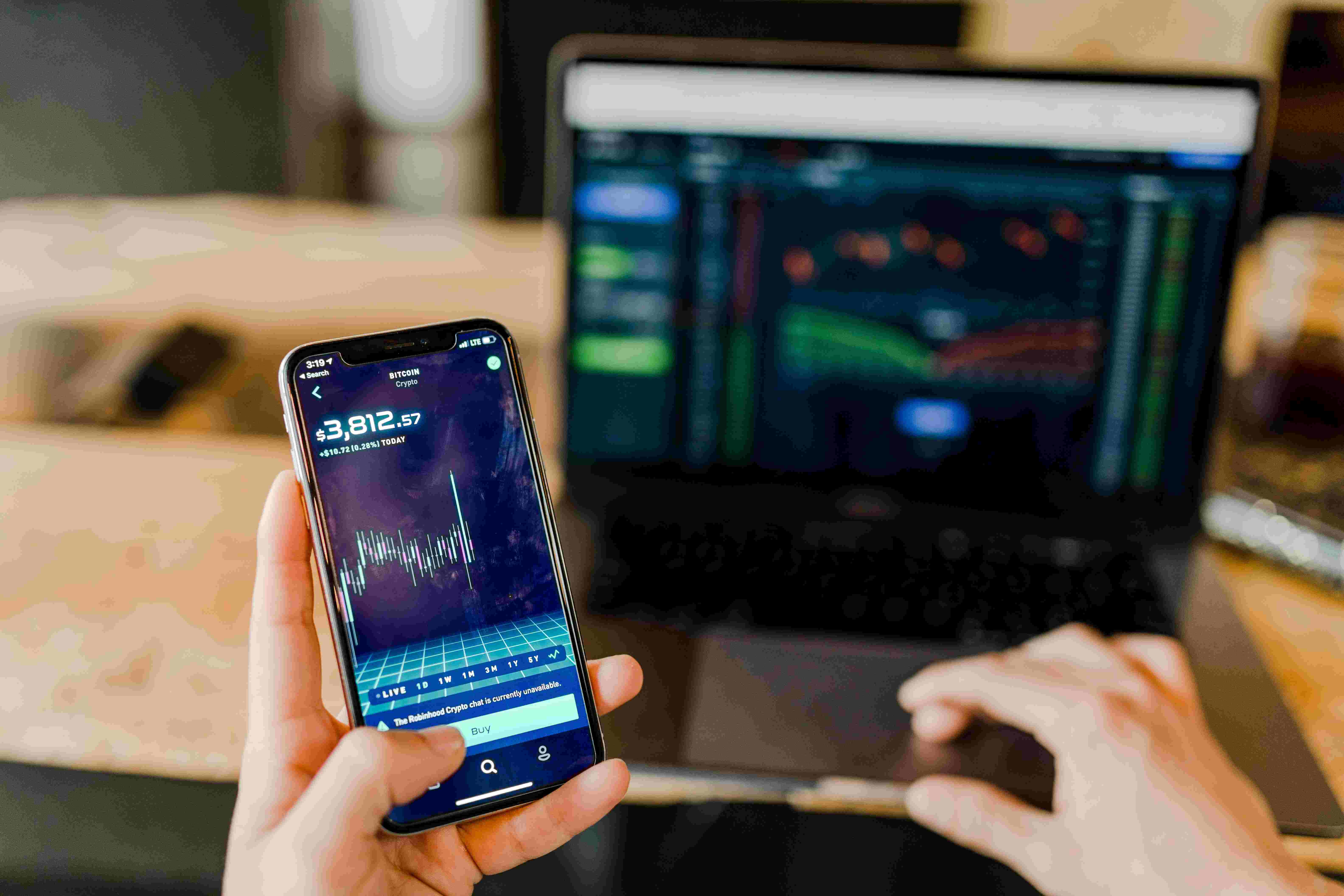 A person holding an iPhone with a stocks balance screen showing on it with a mac laptop in the background showing the market.
