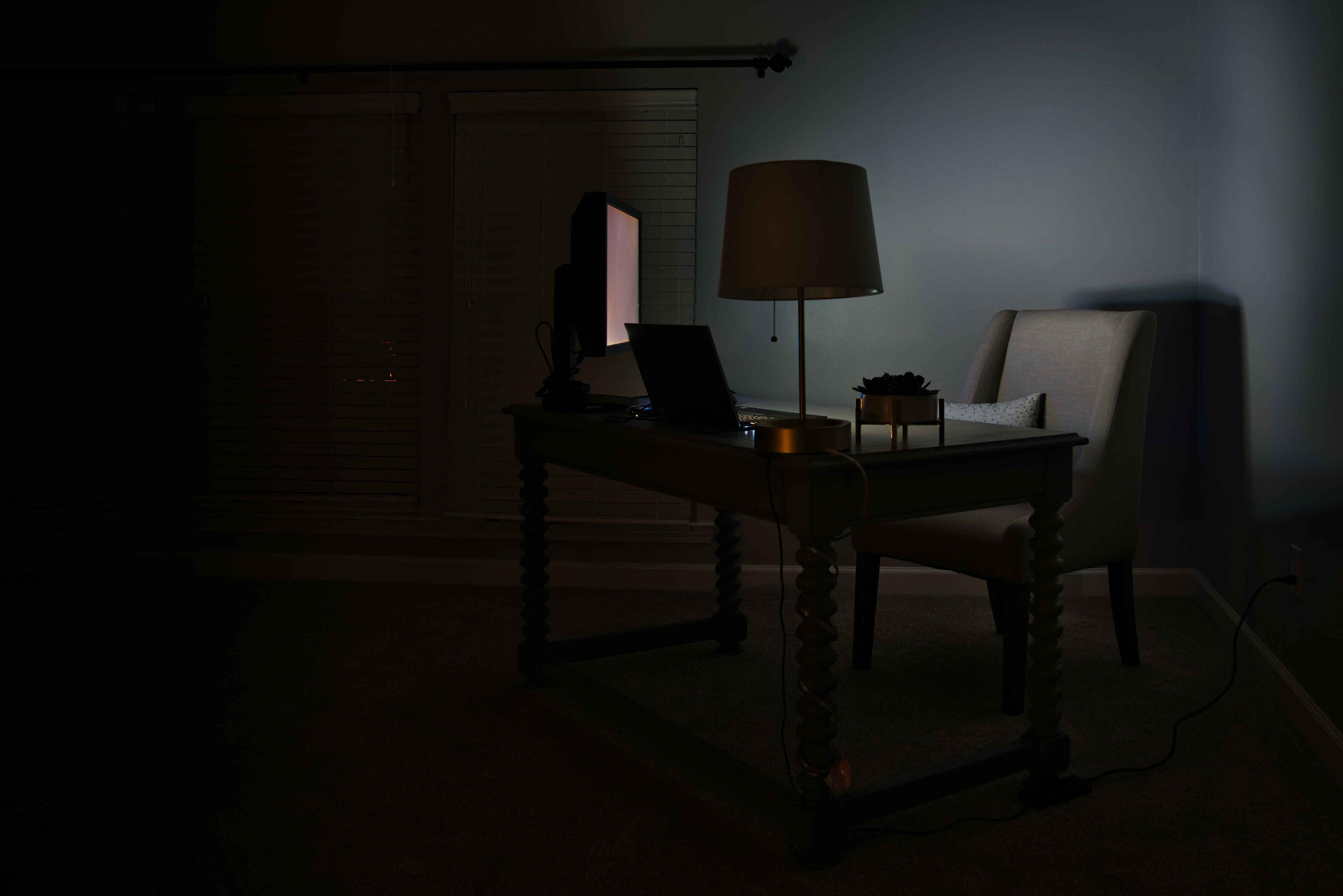 A dark room with a desk that has a plant, laptop, lamp, and a lit-up computer screen on top of it next to a leather chair.