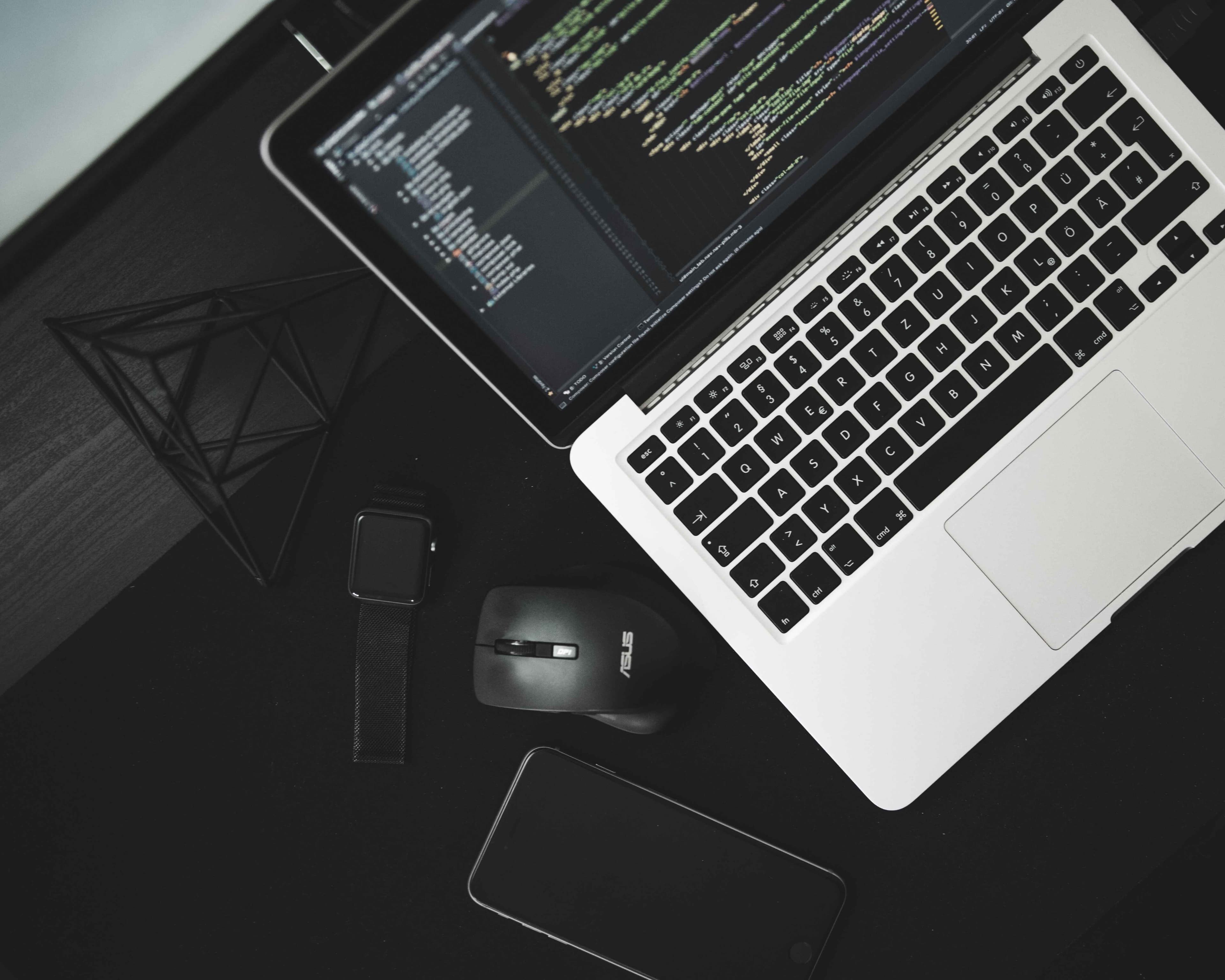 Mac laptop with multiple lines of code shown on its screen next to a black mouse as well as a black iPhone and Apple watch.