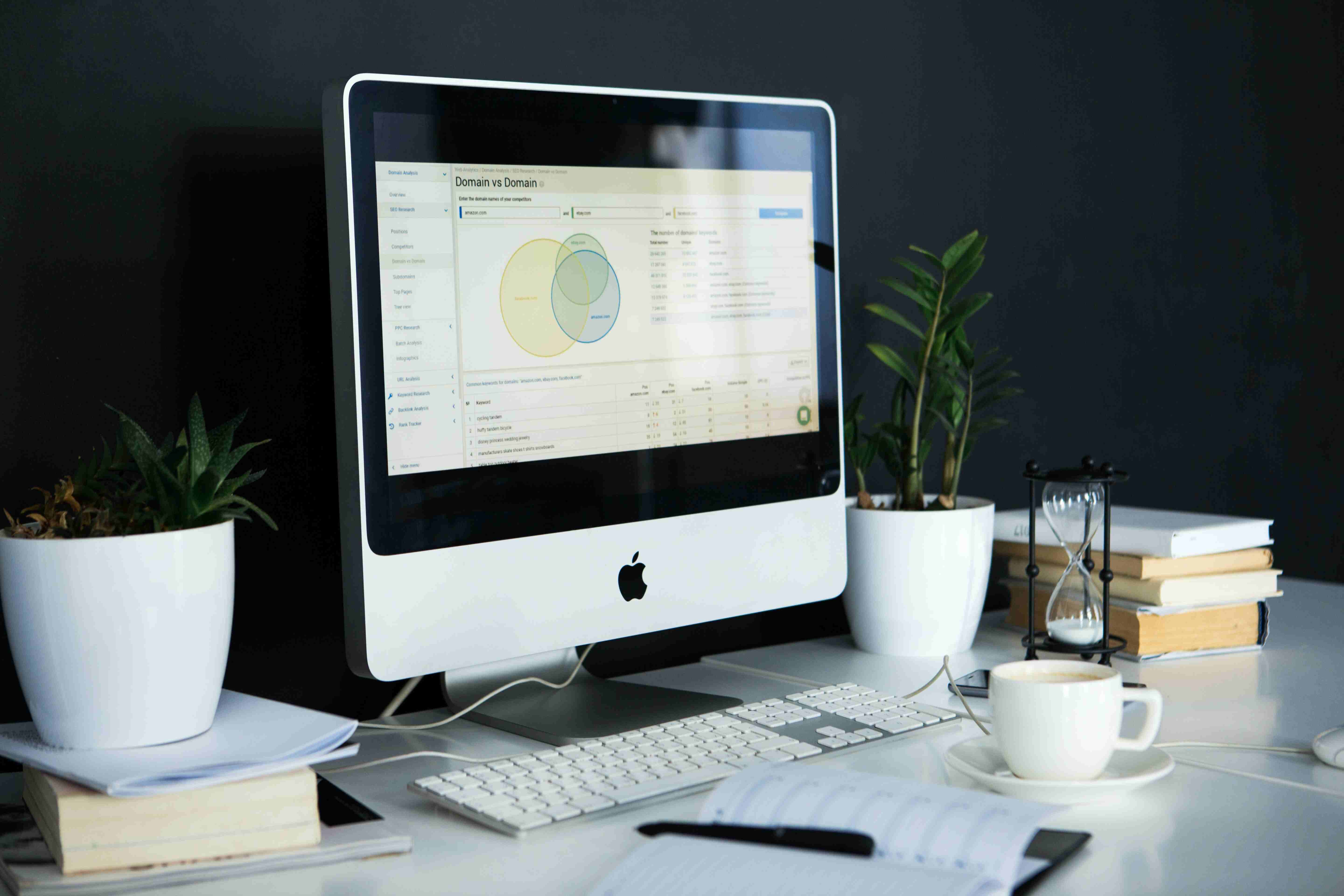 An Apple iMac computer with a keyboard on a white desk with a notebook, pen, two plants, multiple books, and an hourglass.