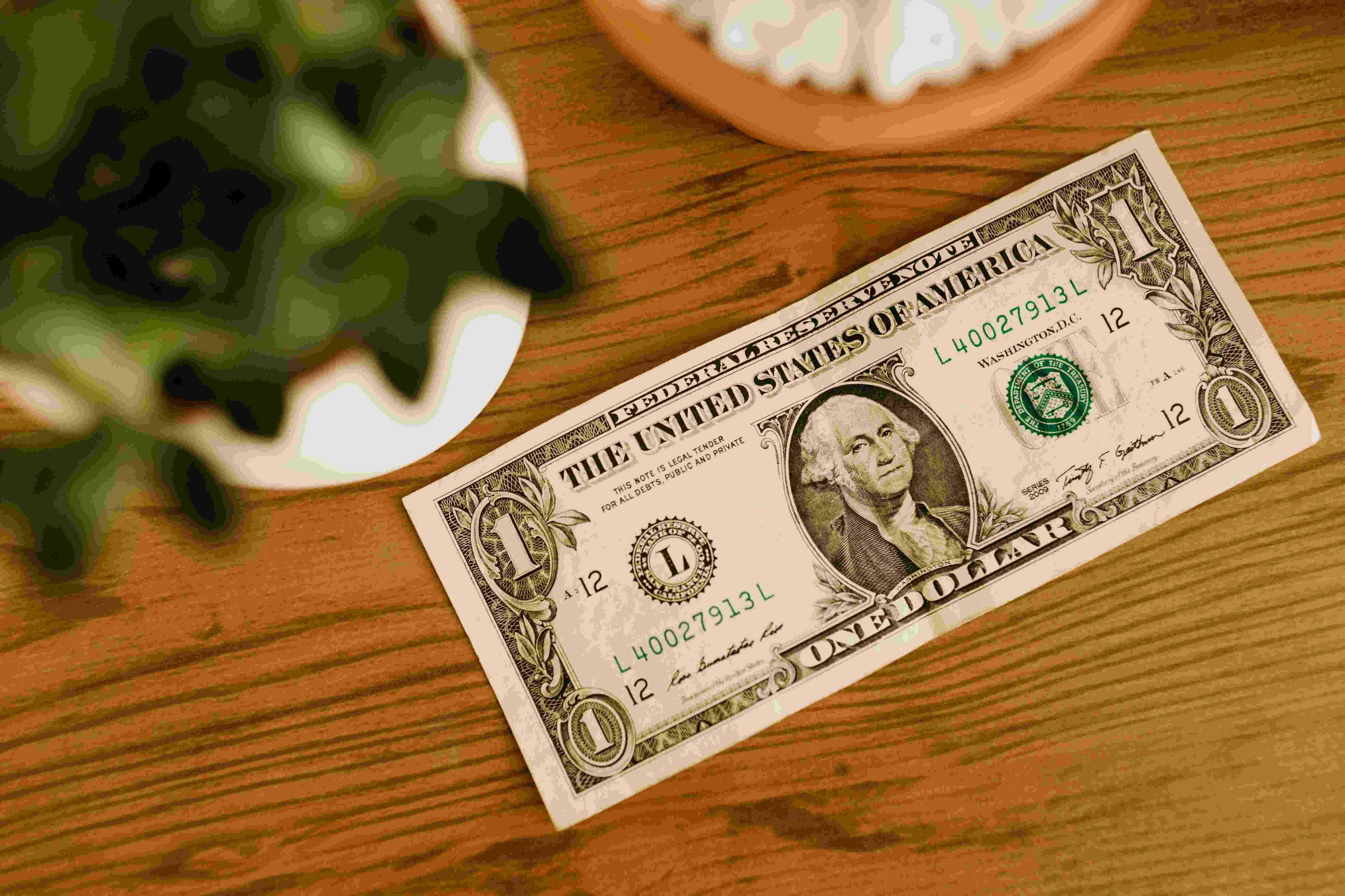A single dollar bill saying face up on a wooden table that has a plant in a white pot on it all next to it and a wooden coaster.