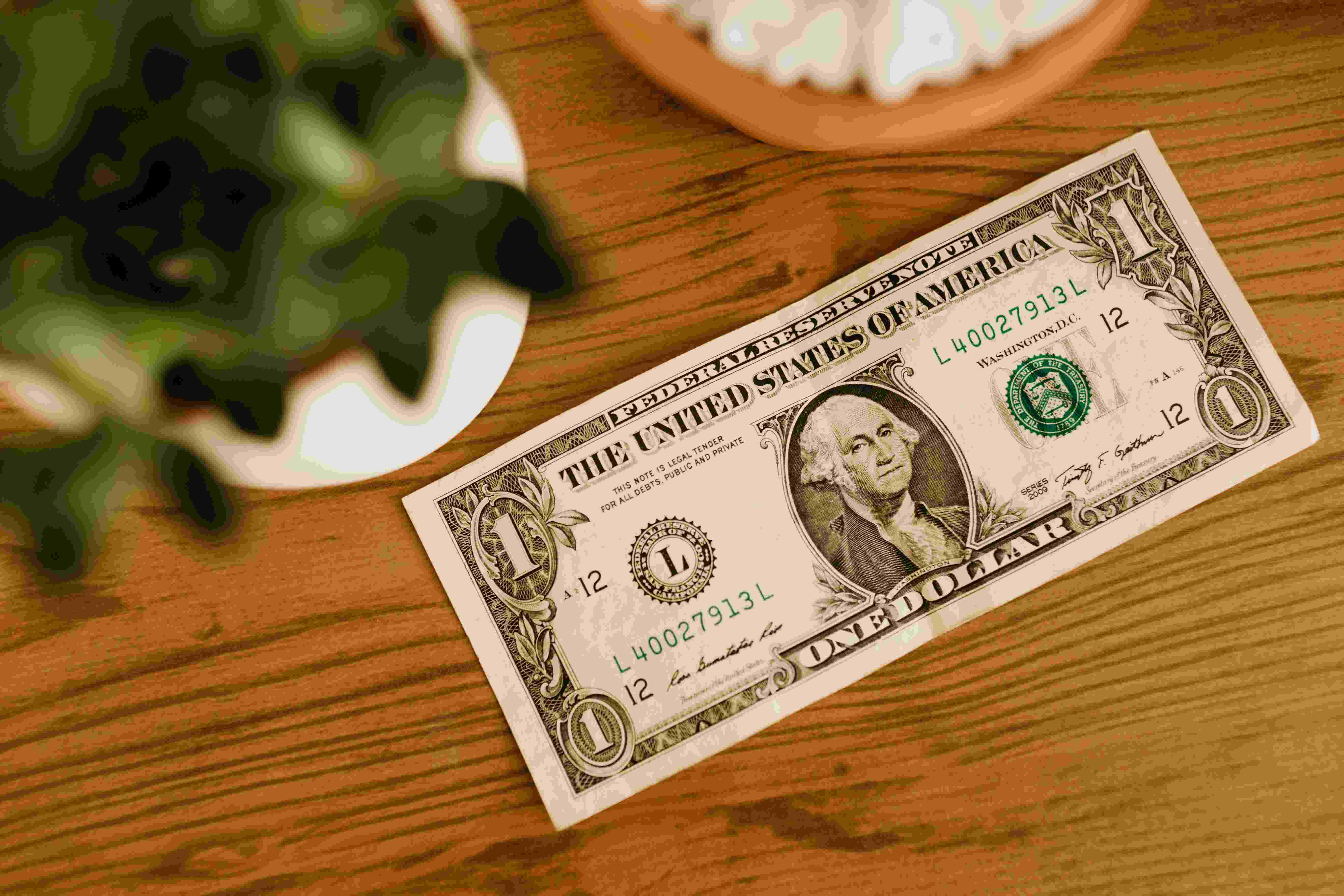 A single dollar bill saying face up on a wooden table that has a plant in a white pot on it all next to it and a wooden coaster.