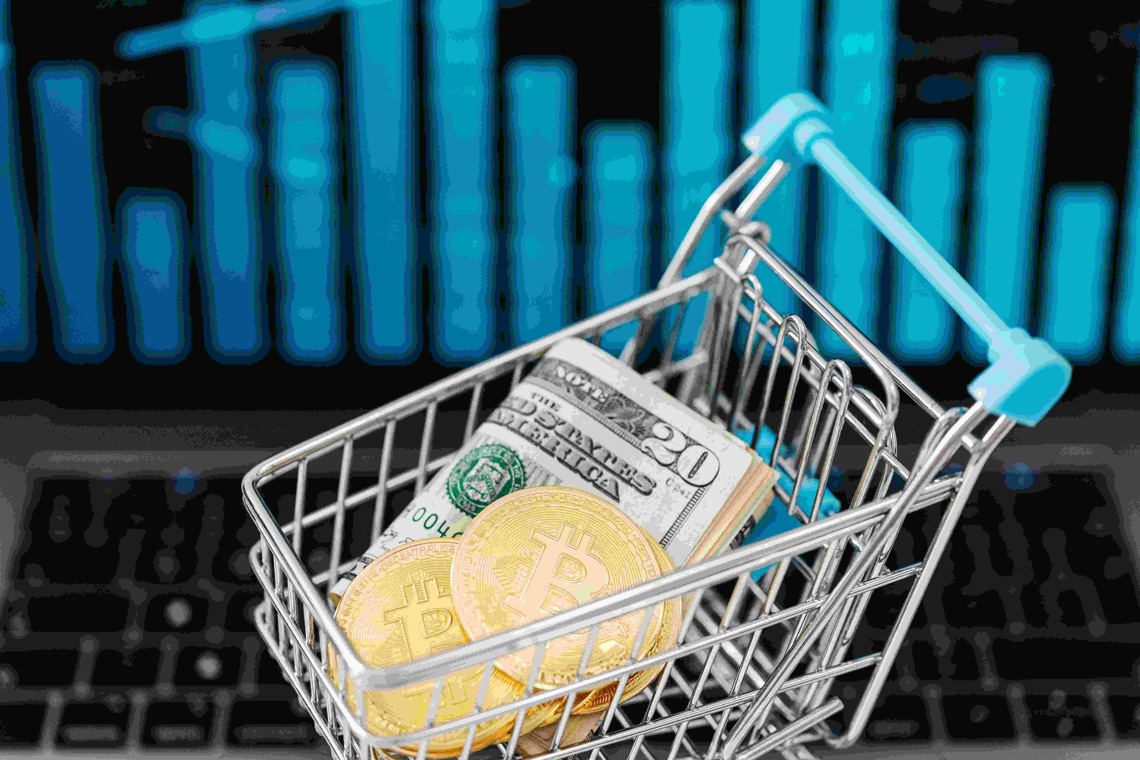 A shopping cart with a couple bills folded up with the twenty showing and two golden coins with the Bitcoin logo all inside it.