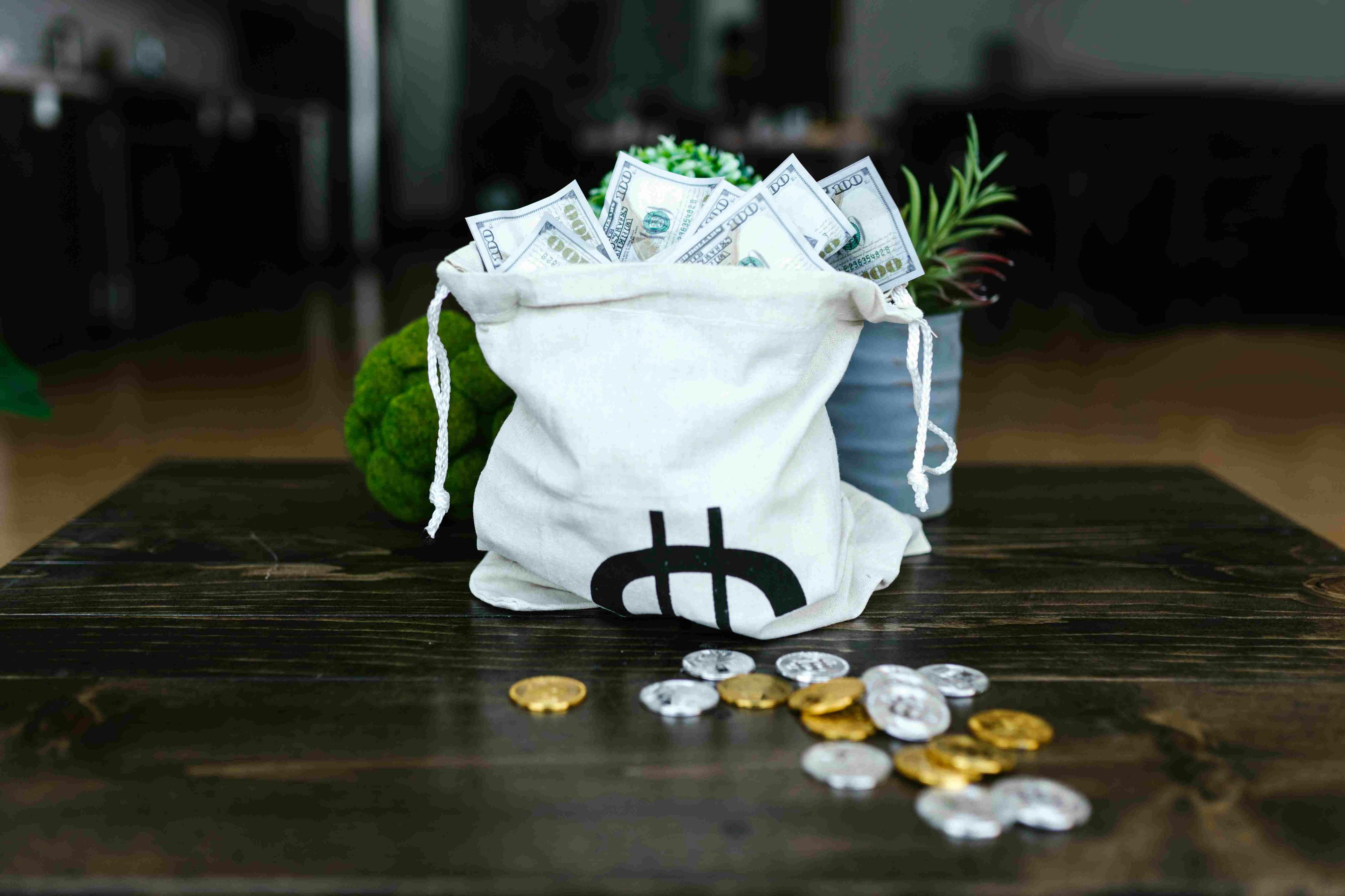 A white money bag filled with stacks of cash next to some gold and silver coin showing the Bitcoin logo all on a brown table.