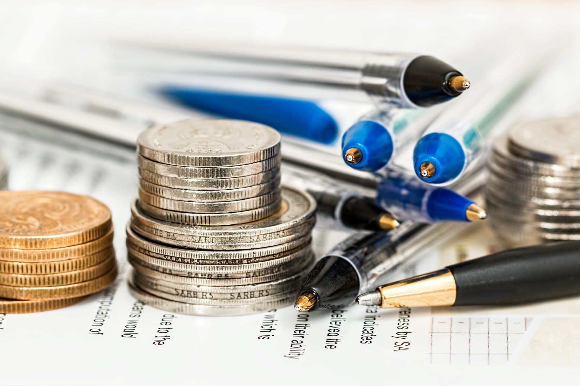 Two stacks of silver coins and one stack of golden coins sitting on top of a piece of paper with writing on it along with pens.