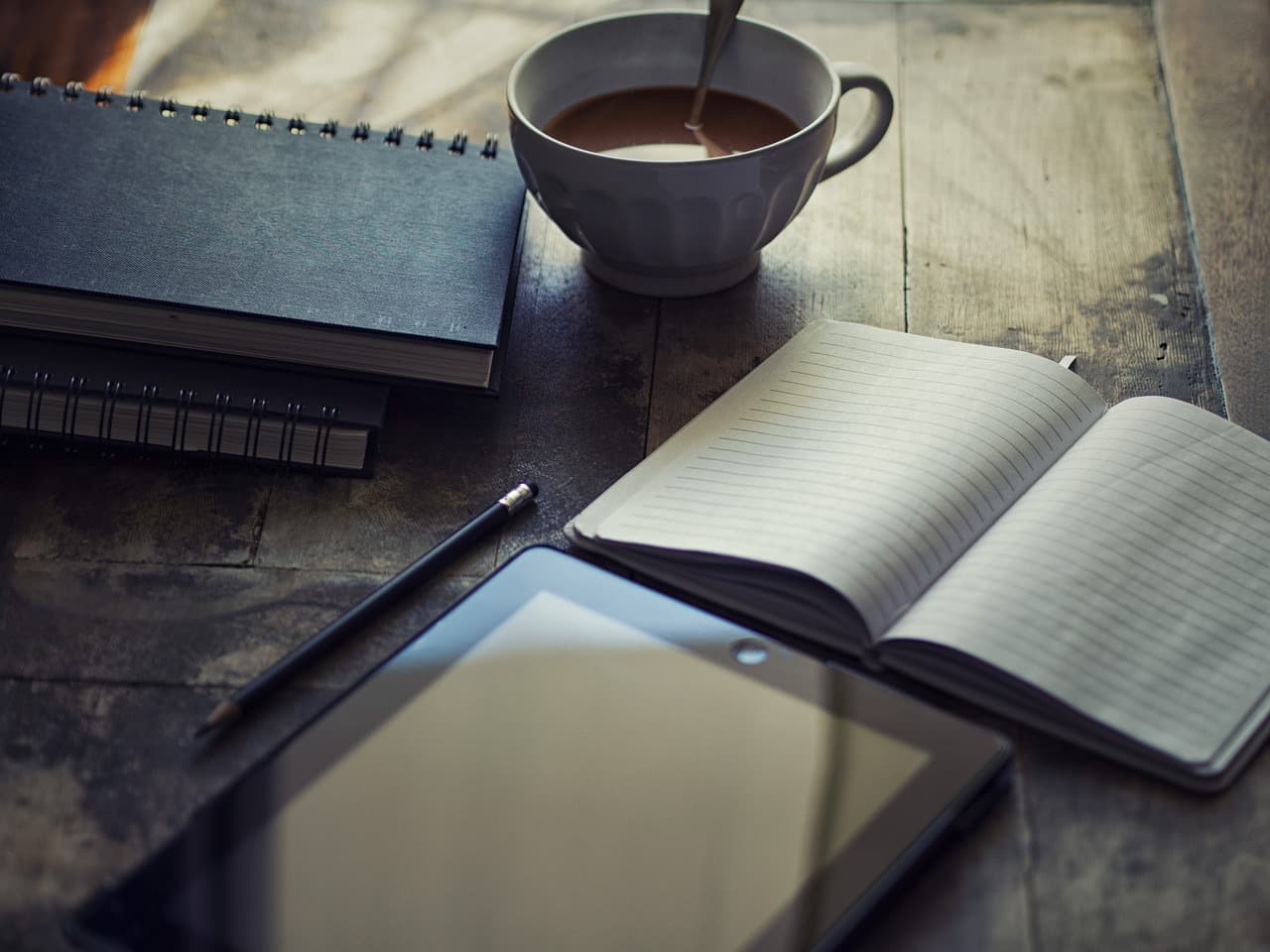 An iPad, an open notebook, two closed notebooks, a pen, and a cup of coffee all sitting on a wooden table in a darkly lit room.