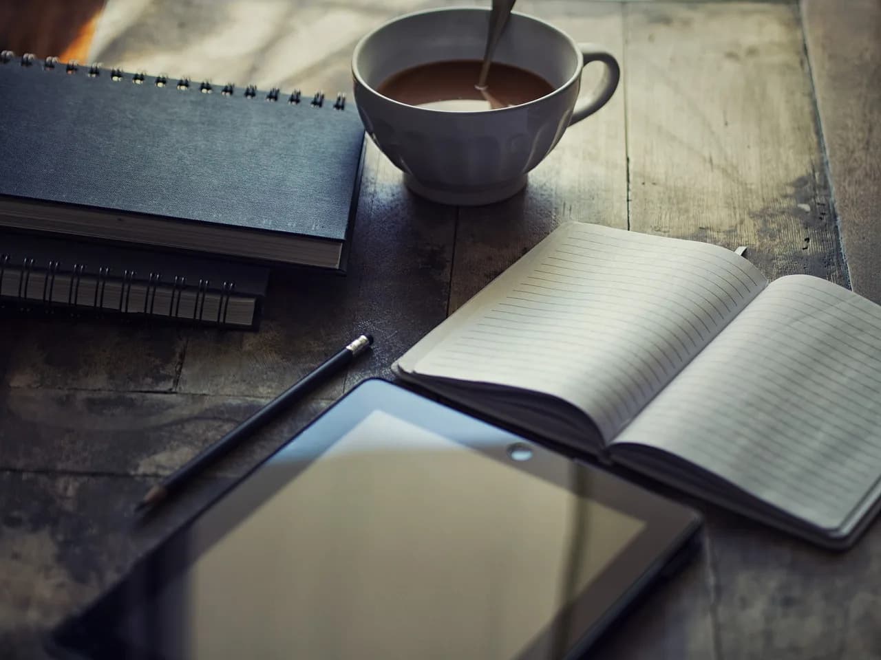 An iPad, an open notebook, two closed notebooks, a pen, and a cup of coffee all sitting on a wooden table in a darkly lit room.
