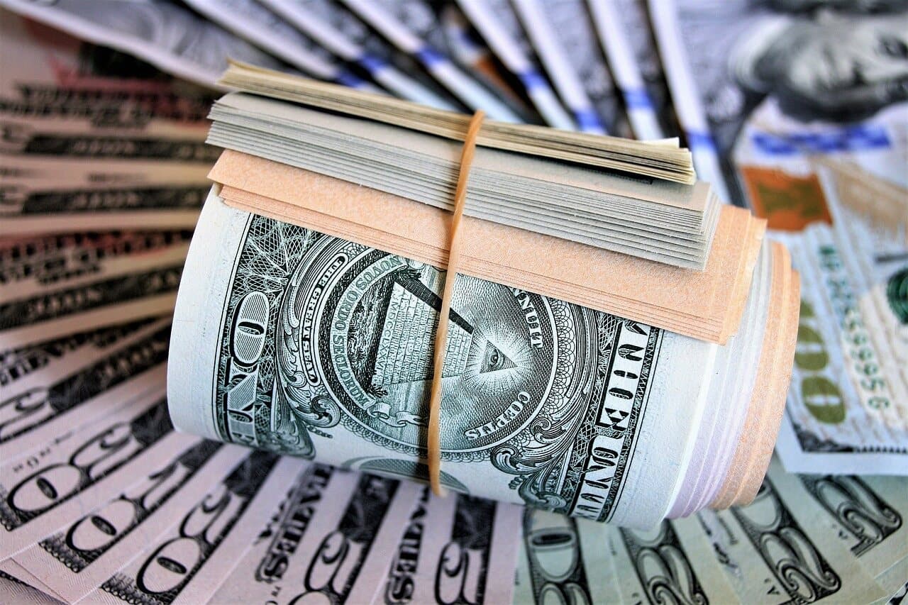 A stack of dollar bills that are rolled up with a rubber band around them sitting on a fan of fifty and hundred dollar bills.