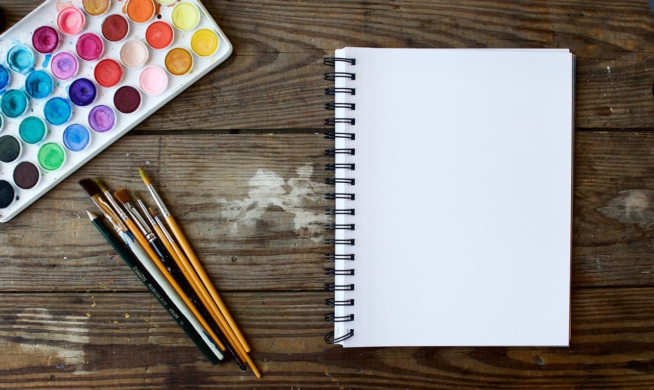 A notebook with multiple paint brushes sitting on a dirty wooden table next to it with a full paint tray of colors beside it.